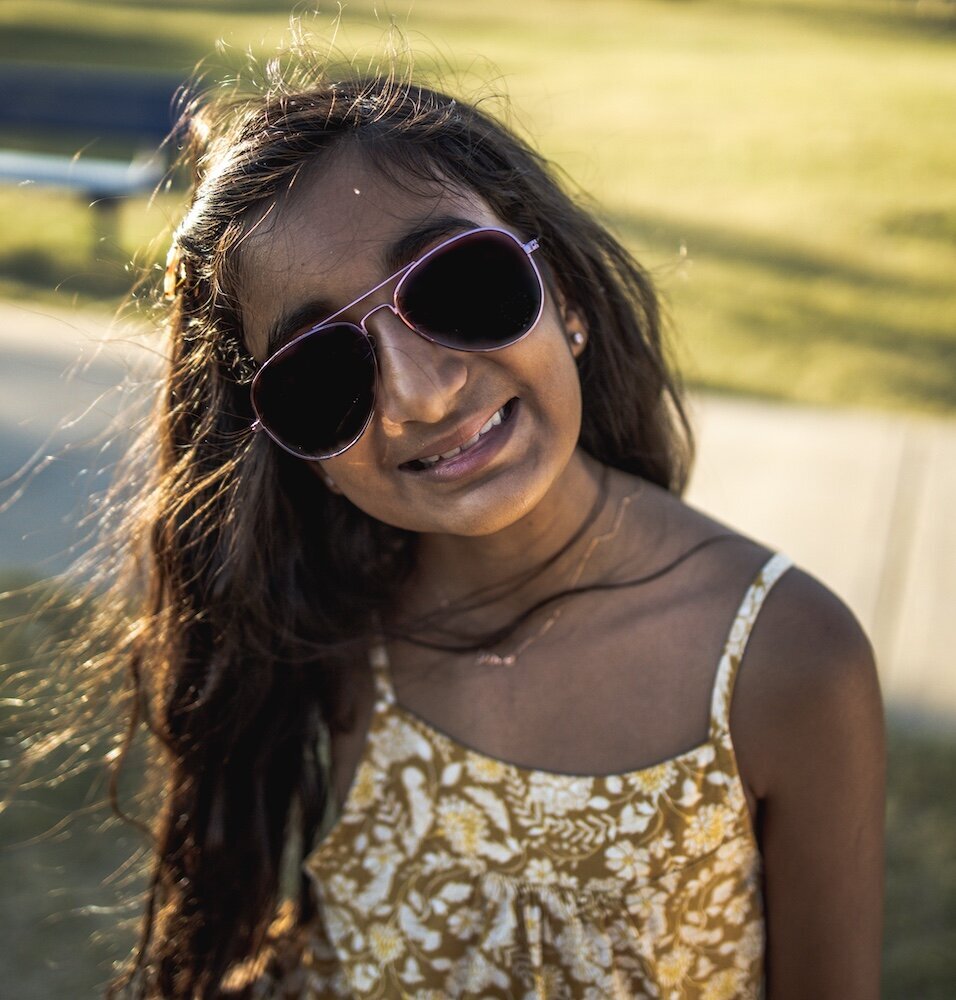 A little girl with long brown hair wearing sunglasses in a yellow floral dress
