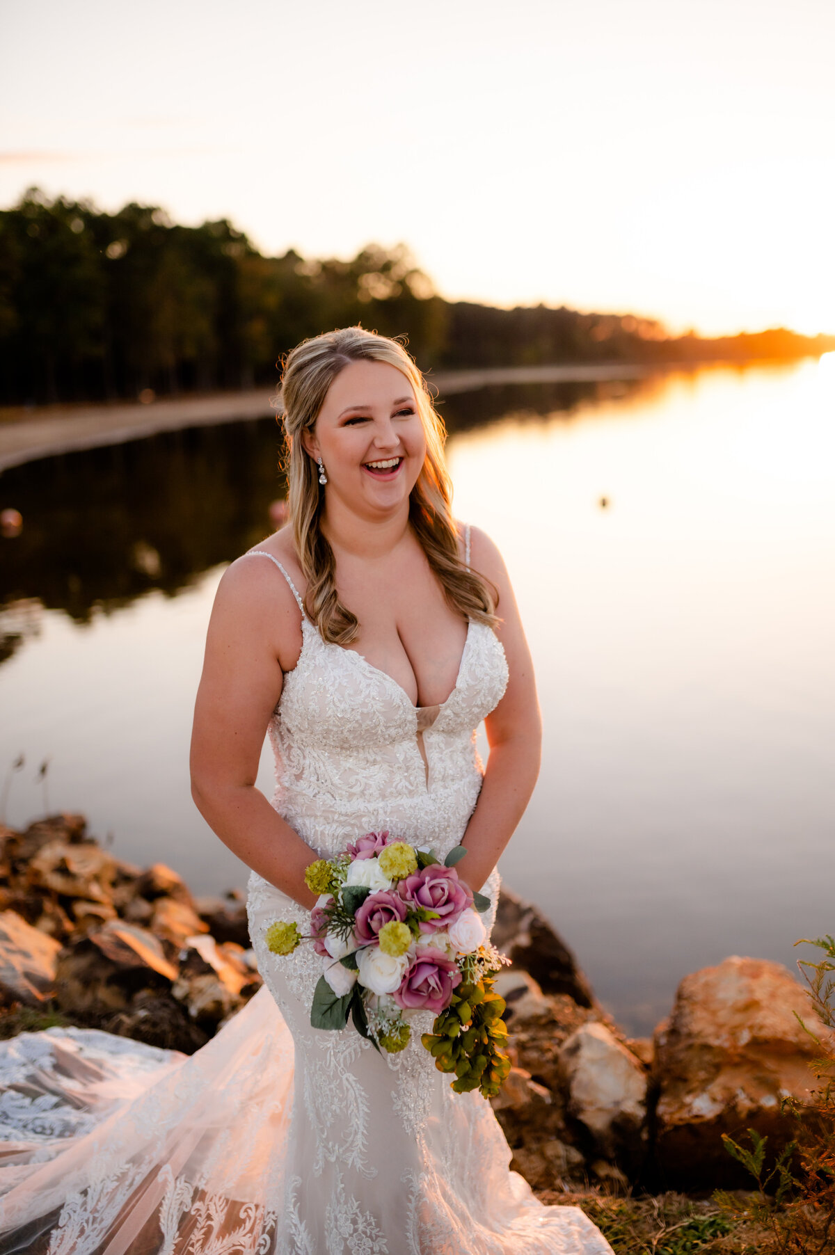 bride laughing as she holds her florals and the sun sets over the lake behind her for her formal bridal session