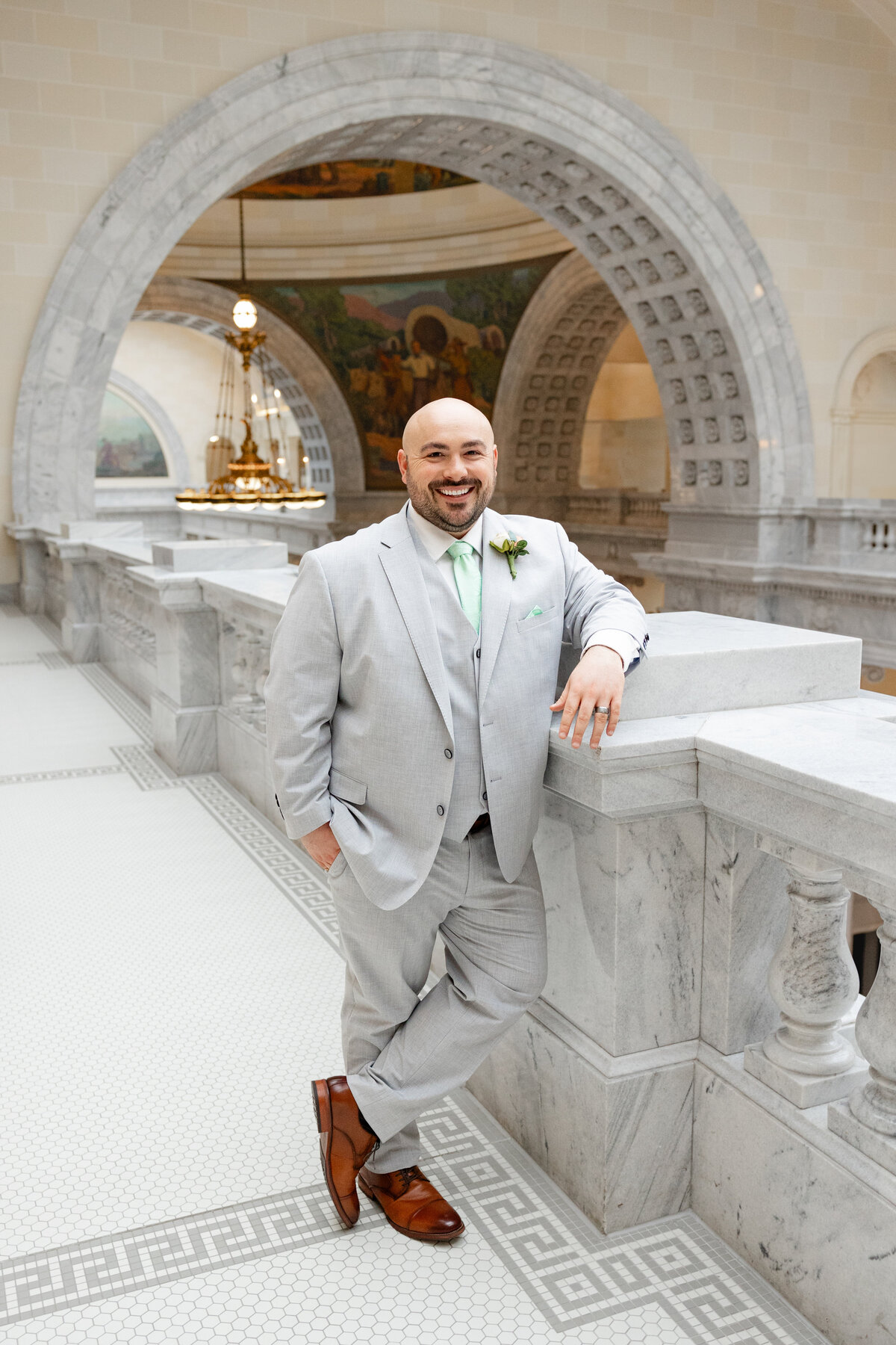 The groom, dressed in a light gray suit with a mint green tie, leans on a marble railing at the Utah State Capitol, smiling confidently.