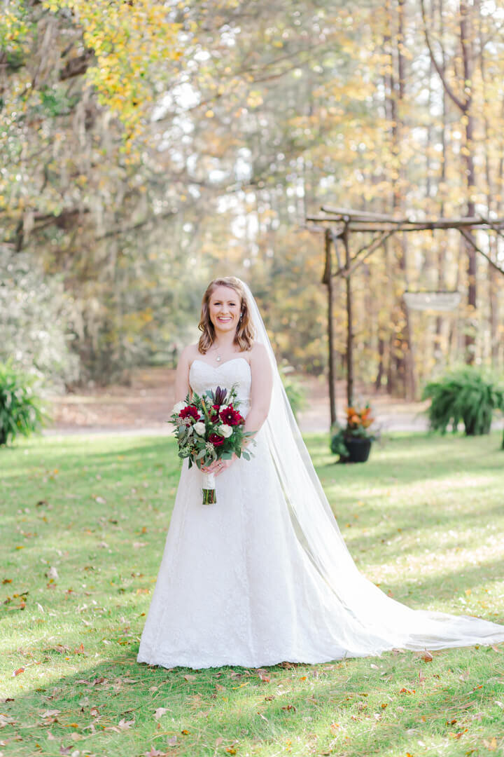 A bride stands with her red, white and green bouquet on a grassy space with trees behind her. Captured by Arkansas wedding photographer Photography by Karla.