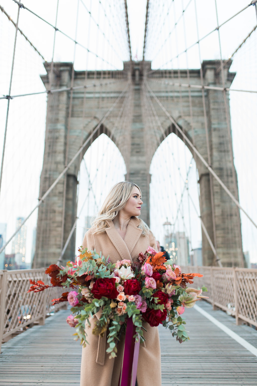 Brittany Frid carries a very large Autumn inspired floral bouquet while standing in front of the Brooklyn Bridge in New York