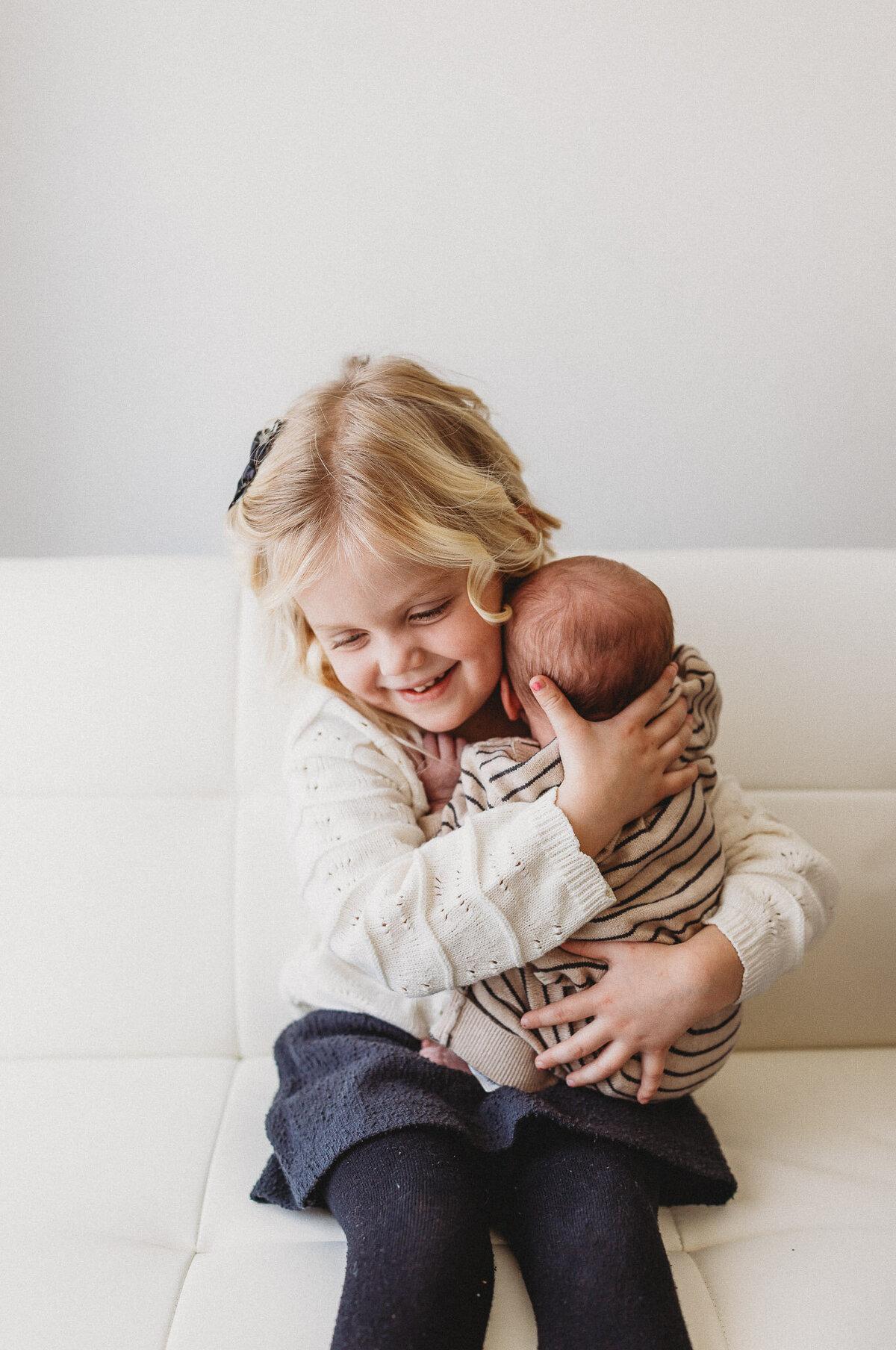 Big sister holding newborn baby brother on her lap on a white couch in white studio setting