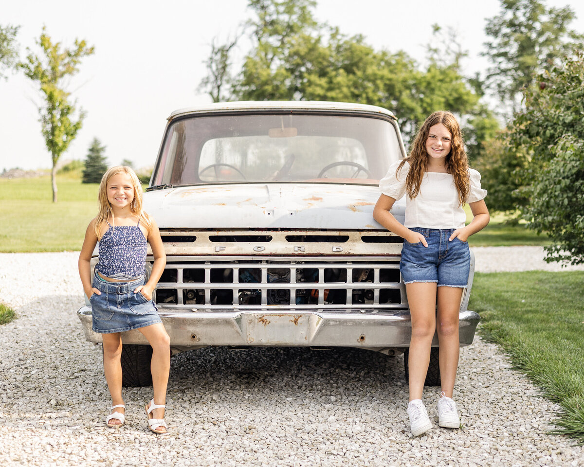 Sisters are standing in front of a classic ford truck in a rocked drive, smiling at the camera. Older daughter is wearing blue jean shorts, a white short sleeve shirt, and has long brown hair.  Youngest daughter has a dark blue and white tank top style shirt with a blue jean skirt, and has medium length blonde hair.