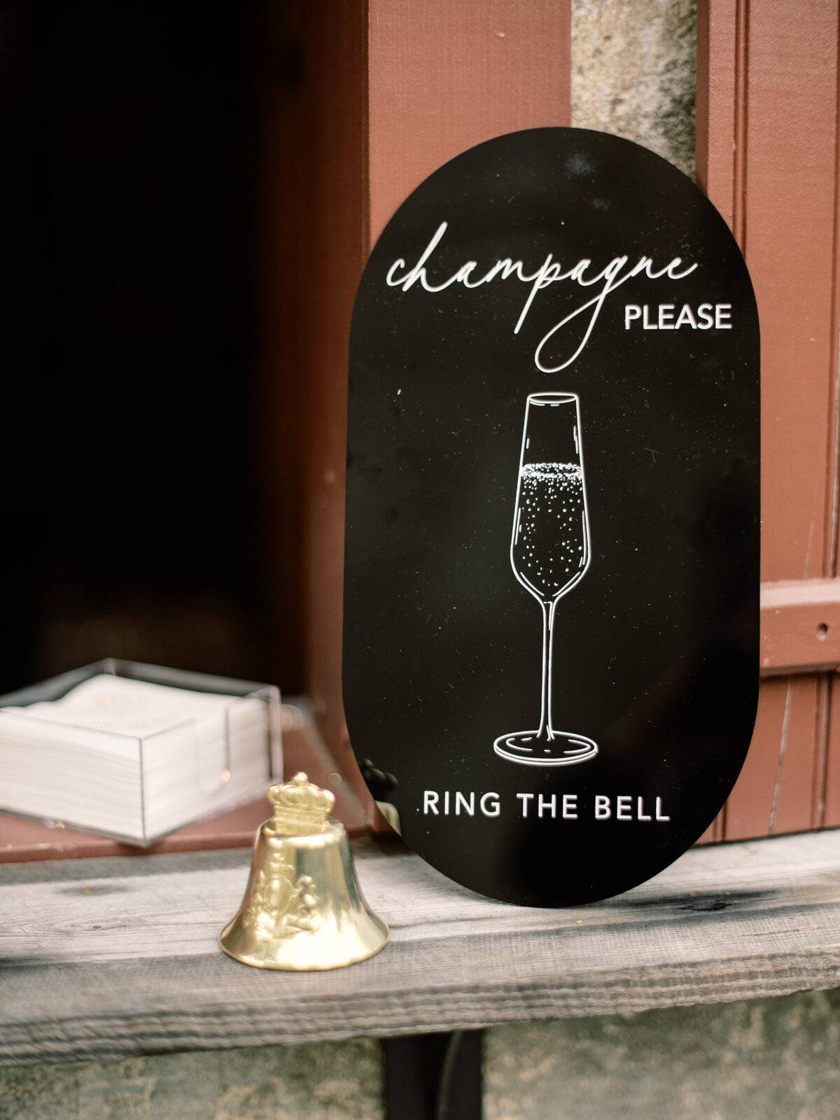 An engaging sign featuring a champagne flute and a bell, inviting guests to ring for celebration drinks, embodying festivity and joyous spirits at weddings.
