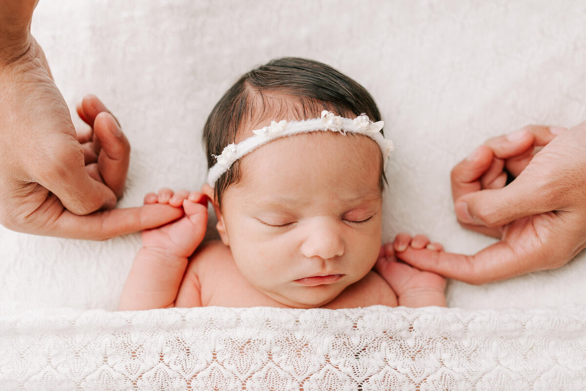 girl laying on white lacy fabric sleeping and holding dad's hands . she is wearing a white headband