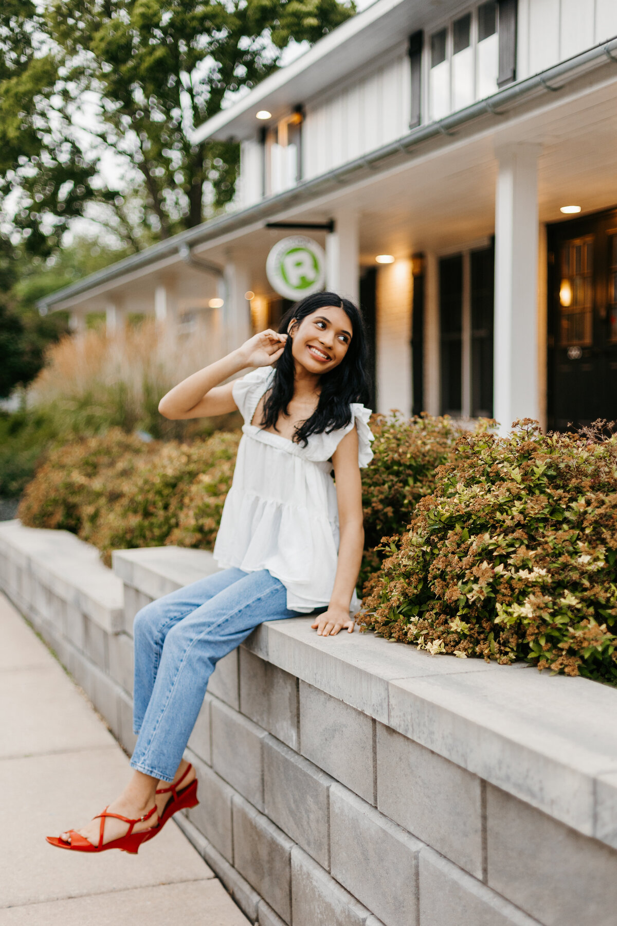 Senior girl in a white top and jeans sitting on a short wall in front of a building and bushes