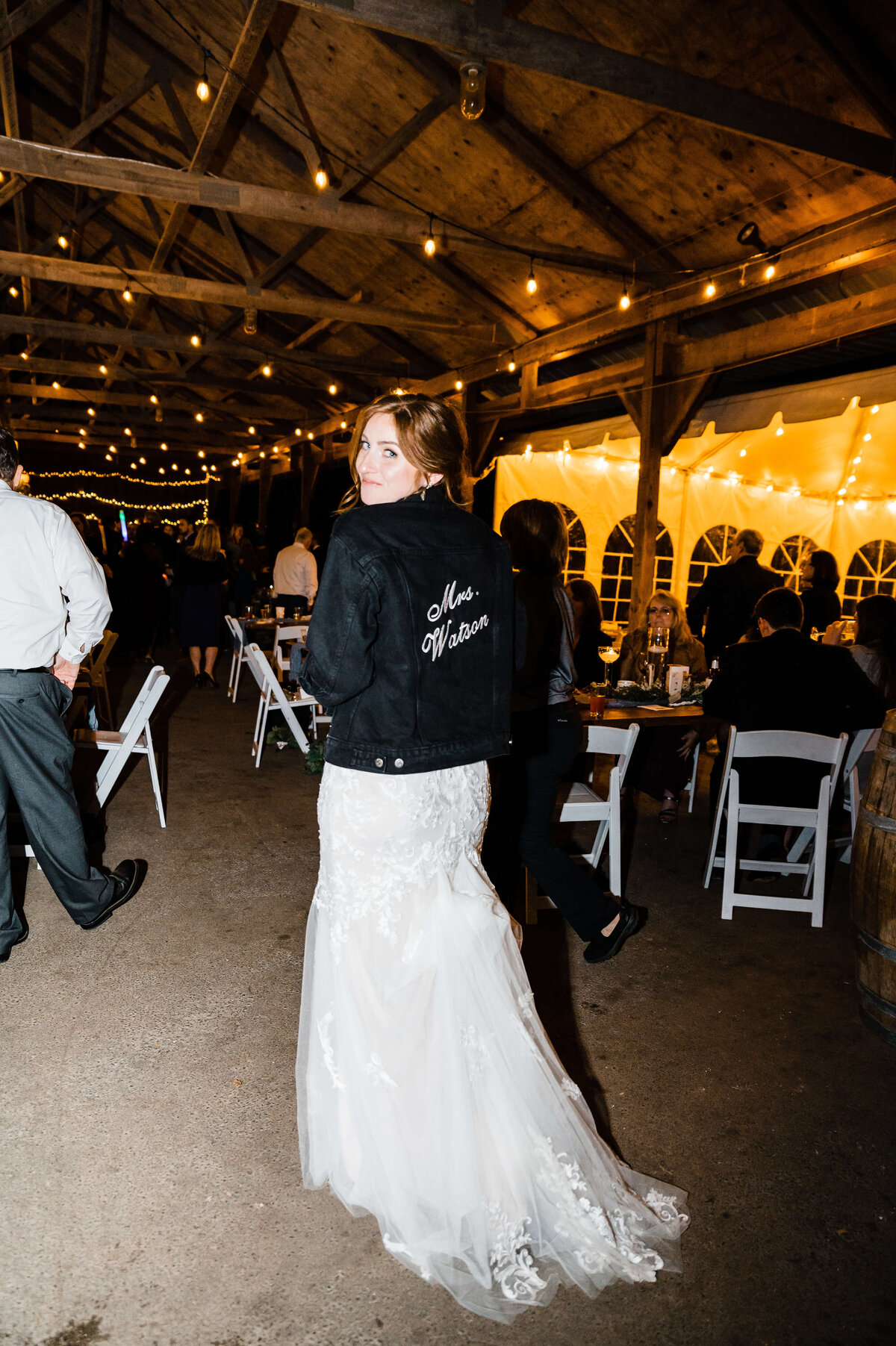 wedding reception in a barn with exposed beams and eddison bulbs strung up in the beams with bride posing in a denim jacket over her gown and smiling over her should to show off her last name on the back of the jacket