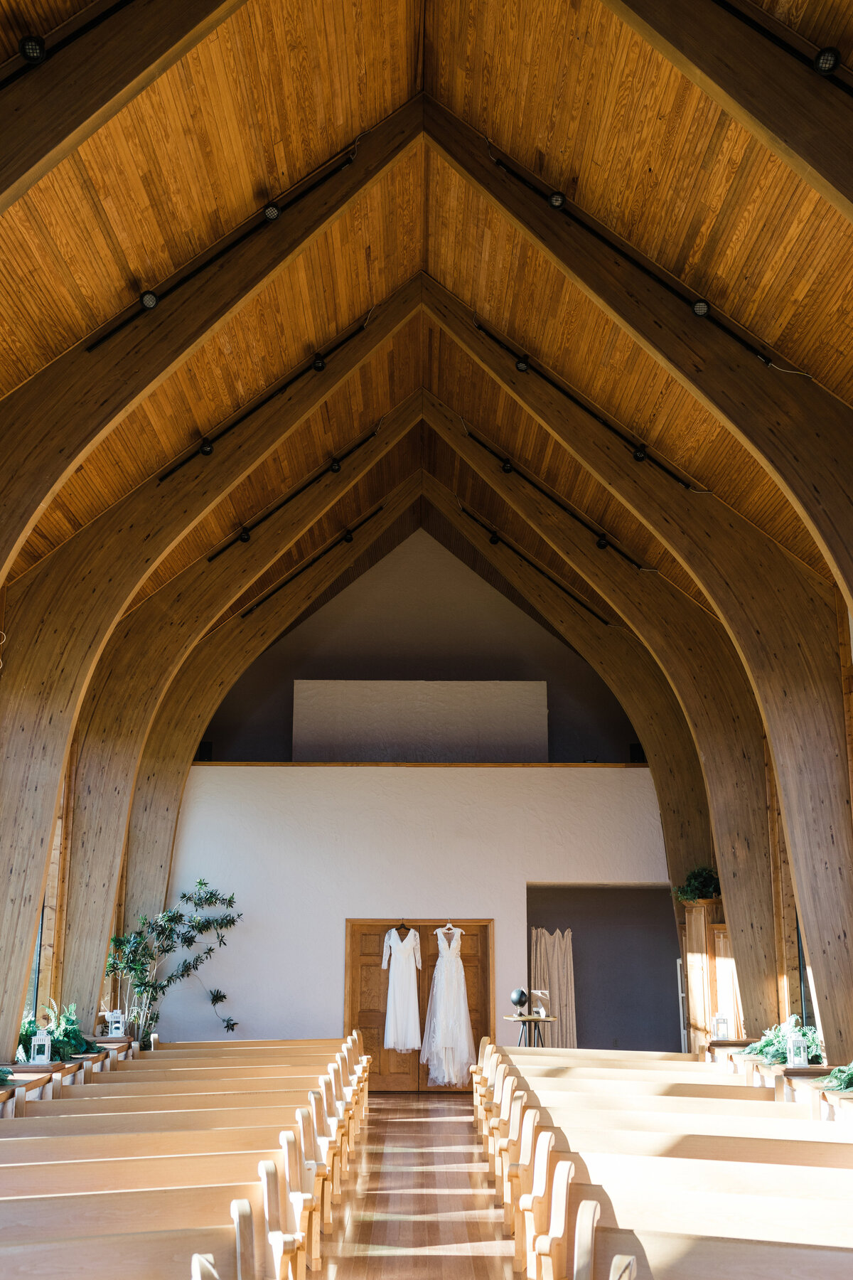 Detail shot of two wedding dresses hanging next to each other in a large, beautiful chapel. The dresses are both white with one being short with sleeves and the other being long and flowing without sleeves. The chapel has a large flowing, curved, wooden ceiling with many pews and flowers in the foreground.