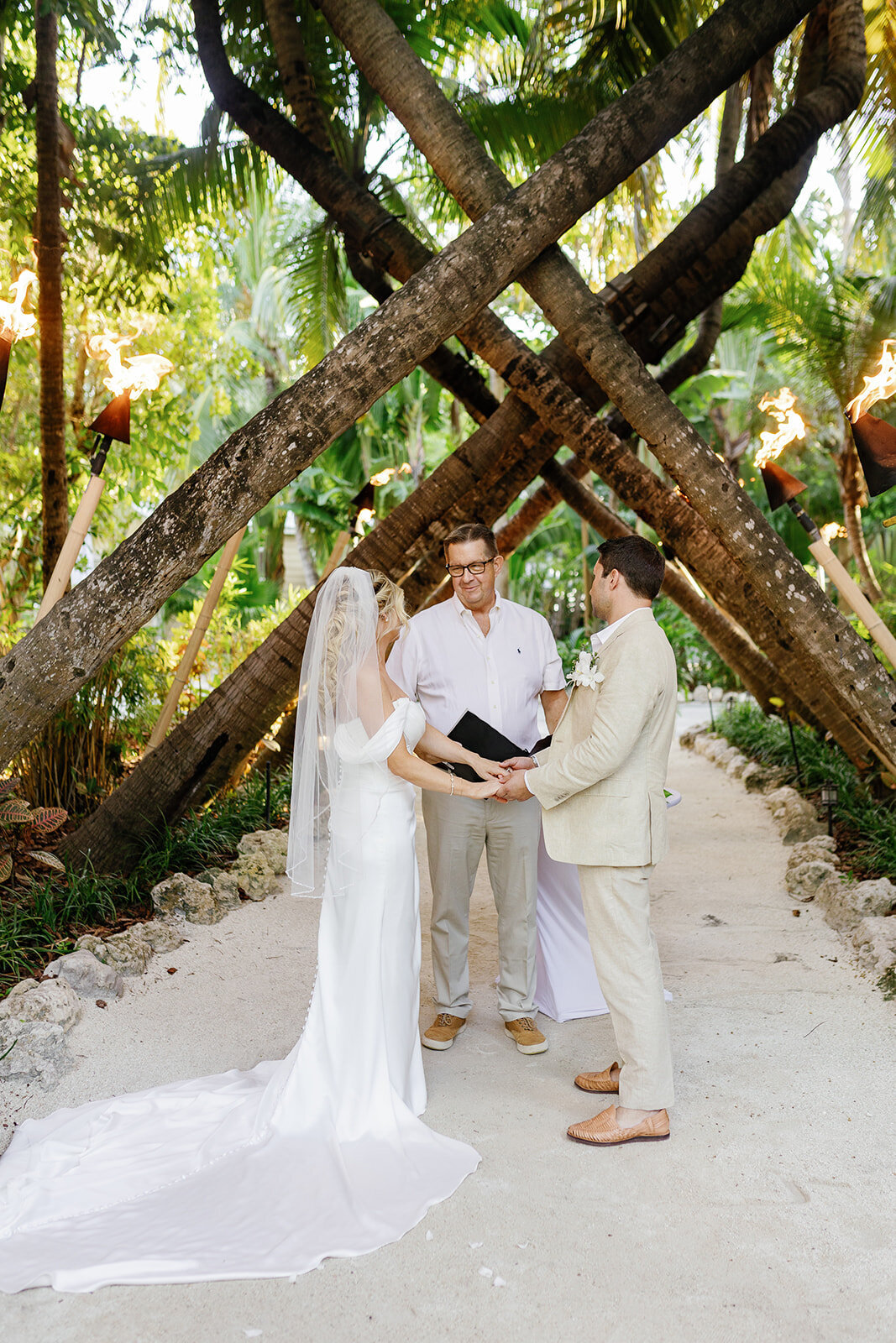 Monica and Austyn stand under a wooden arch with tiki torches, saying their vows at their intimate Islamorada wedding.