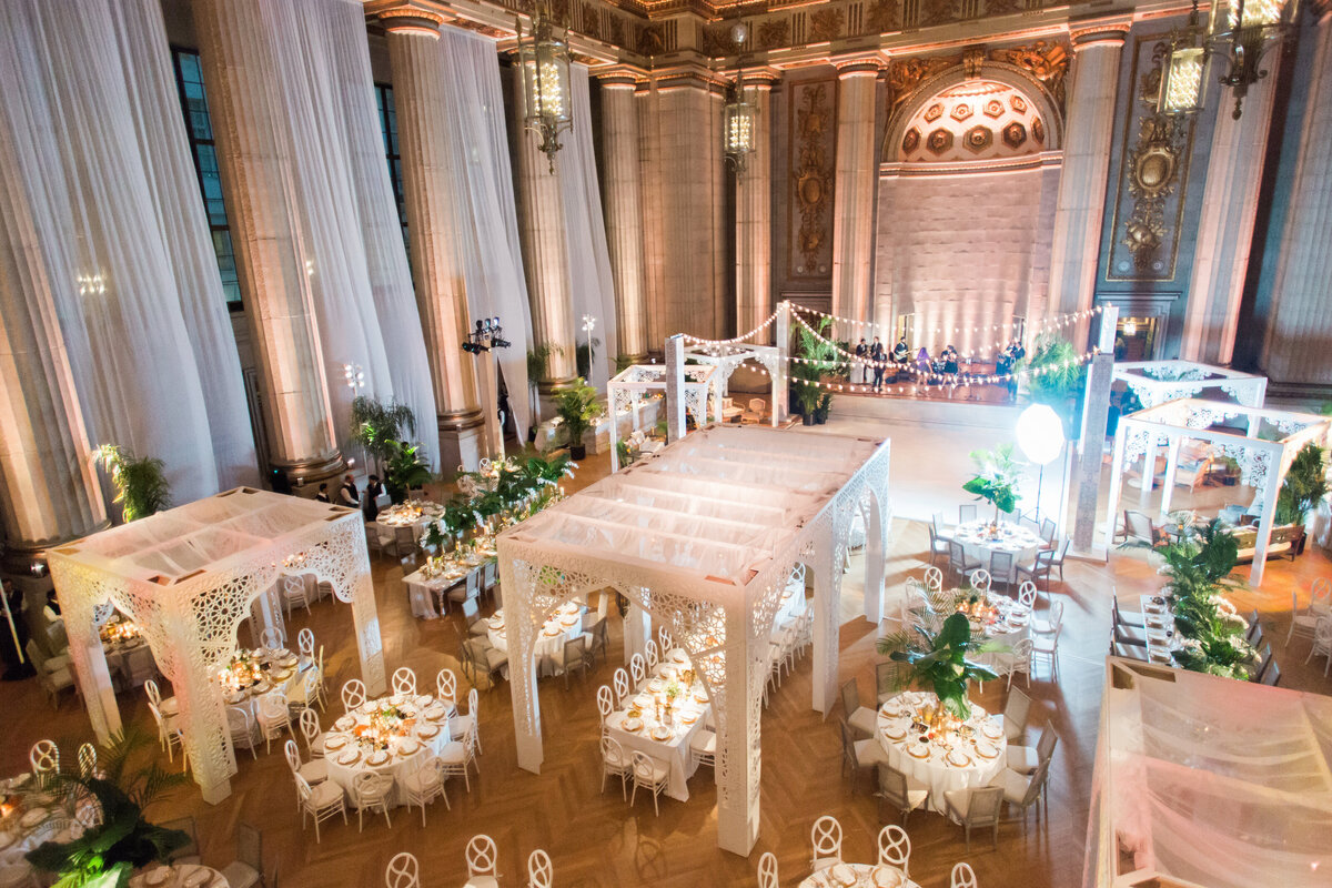 The photo was taken at the Mellon Auditorium in Washington D.C., which is known for being one of the best luxury DC venues with European flair. It was captured by photographer Sarah Bradshaw. The elegant wedding reception in the grand hall features high ceilings, ornate columns, and draped white fabrics. The tables are beautifully set with fine china and are surrounded by white chairs. White pergolas with string lights create a romantic ambiance, while lush green plants complete the decor, adding a touch of natural beauty to the stunning setting.