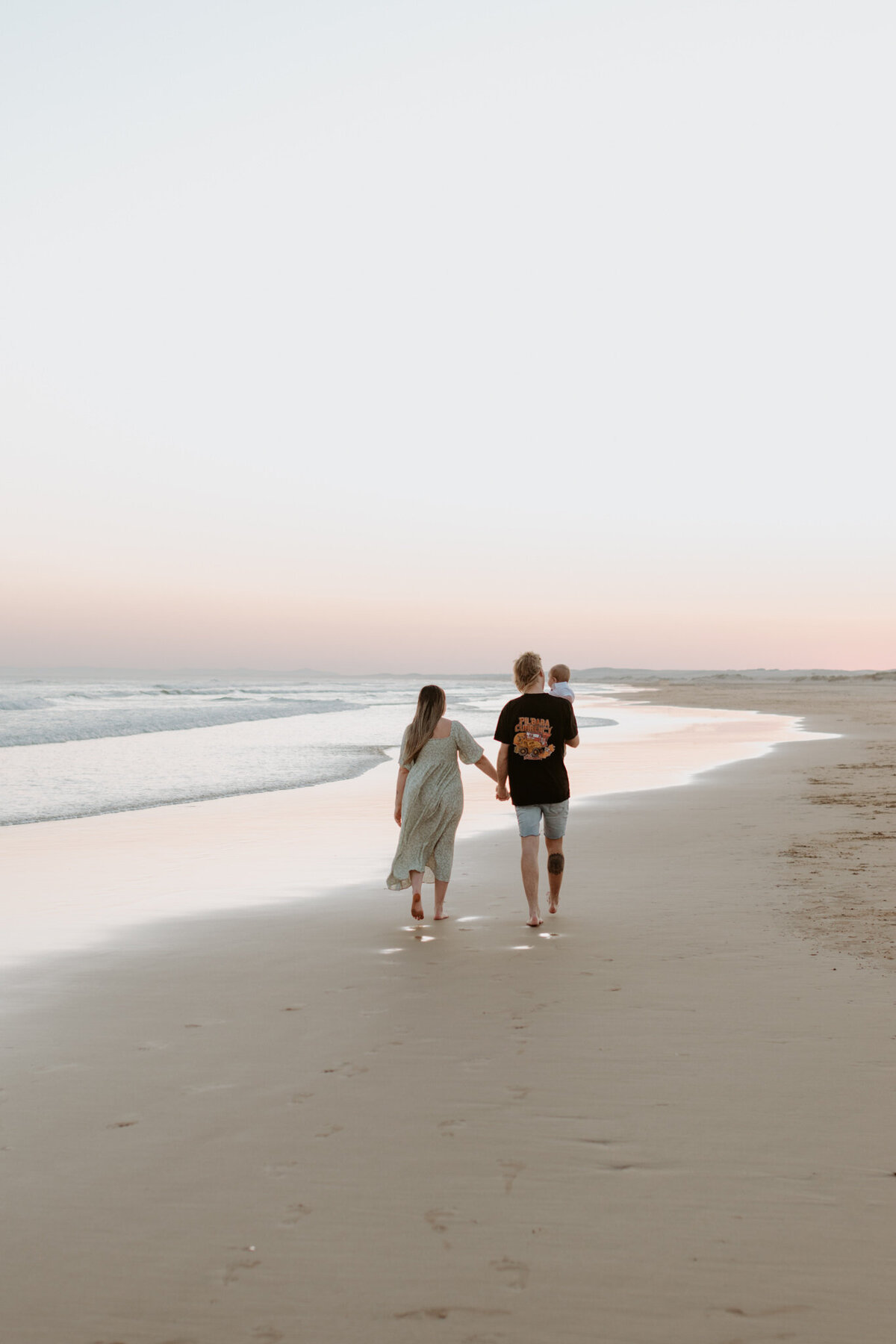 Young family walking hand-in-hand along the shoreline at sunset, with soft pastel skies and footprints in the sand during a relaxed beach photography session.