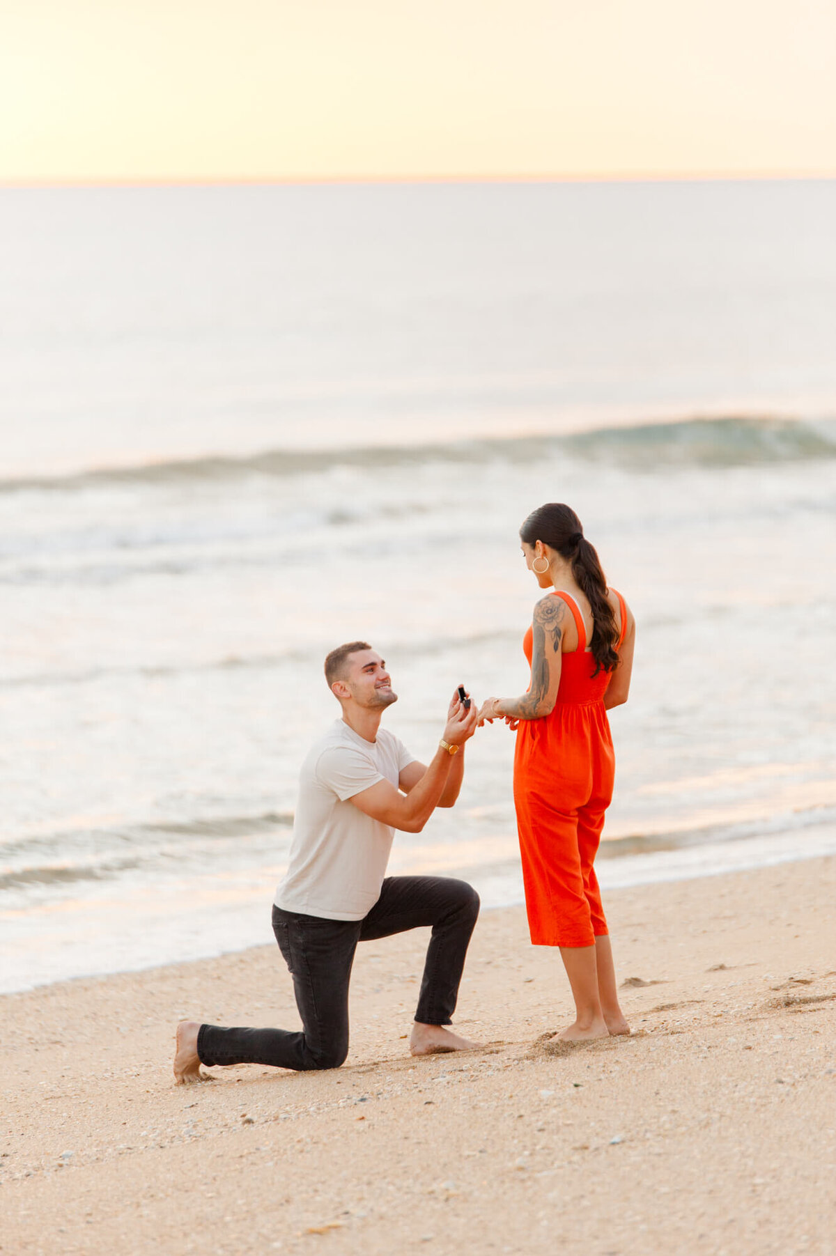 Couple stands at the shoreline with boyfriend on one knee proposing to his beautiful girlfriend at sunrise