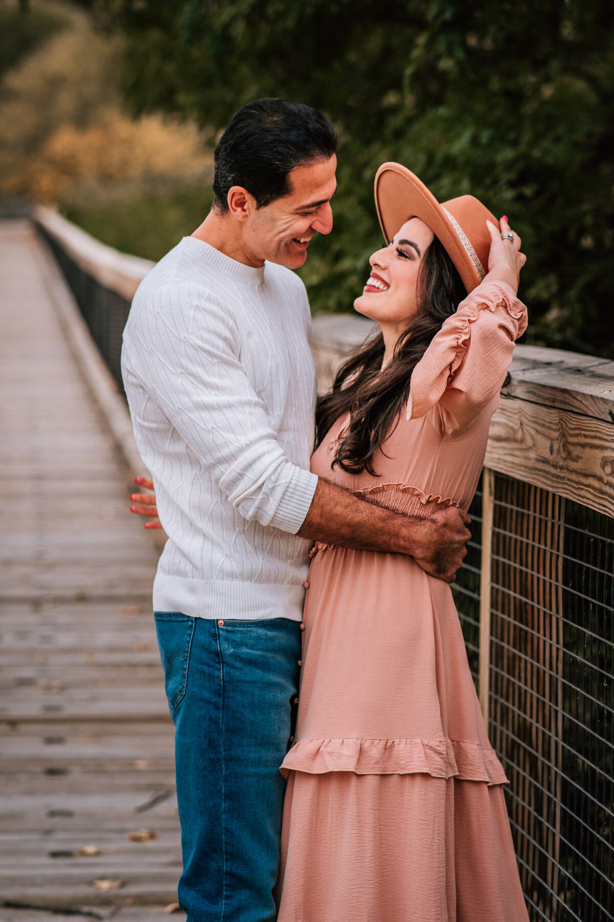 A color photo of a couple standing on a wooden bridge. The woman is wearing a wide-brimmed hat and a flowing dress, while the man holds her waist, both smiling joyfully at each other.