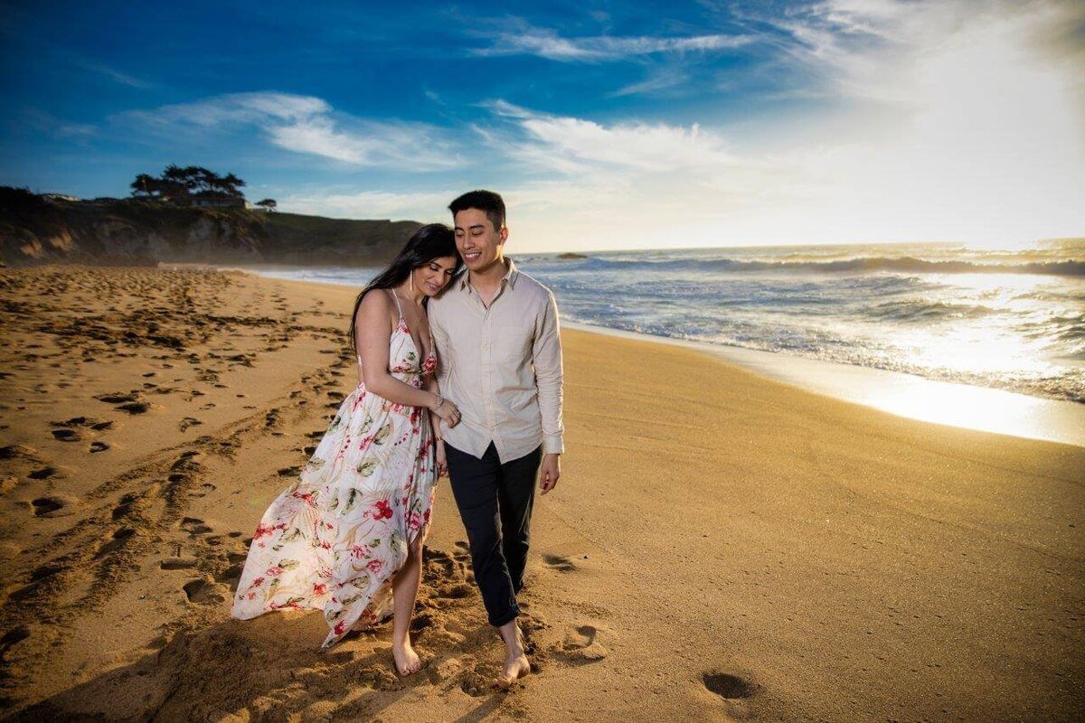 Engaged coupel walks on the beach  with the woman in a flower long dress with her head on the man's shoulder as they hold hands and walk. Photo captured by sacramento wedding photographer philippe studio pro.