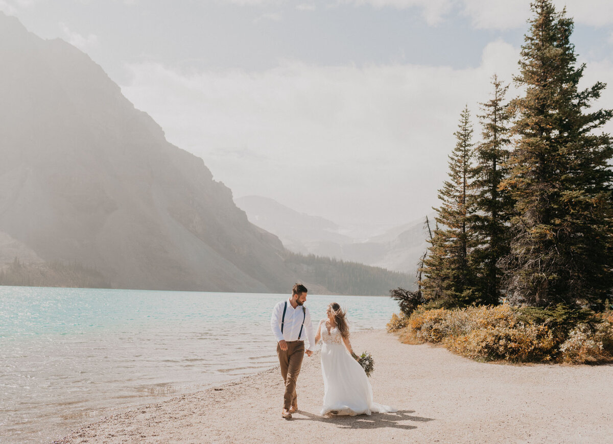 Bride and groom holding hands, walking along a beach in the mountains of Alberta, captured by Ash Maclean Photography, romantic elopement and wedding photographer in Red Deer, Alberta. Featured on the Bronte Bride Vendor Guide.