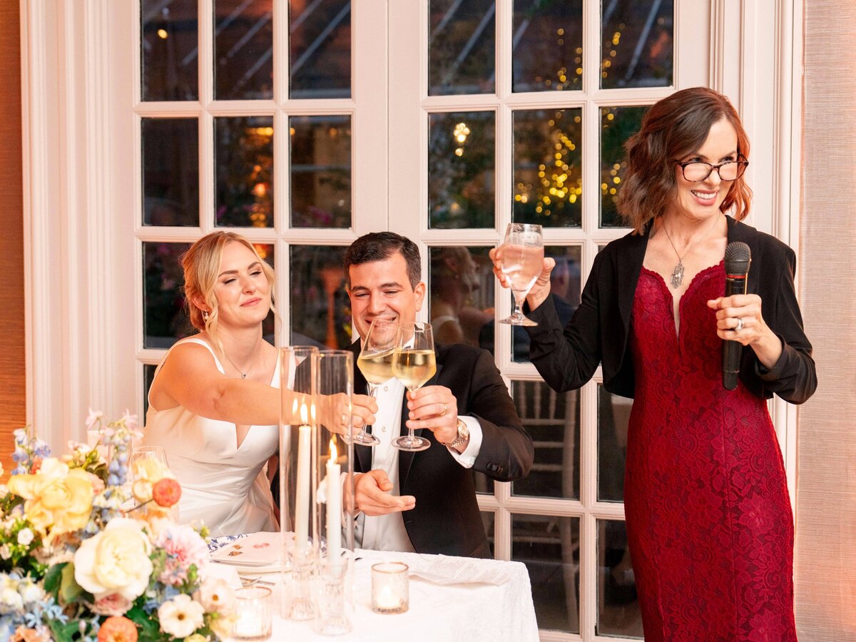 A woman in a white dress and a man in a suit sit at a table, raising glasses for a toast. A woman in a red dress stands next to them, holding a microphone and a glass. They are in a room with large windows and floral arrangements on the table.