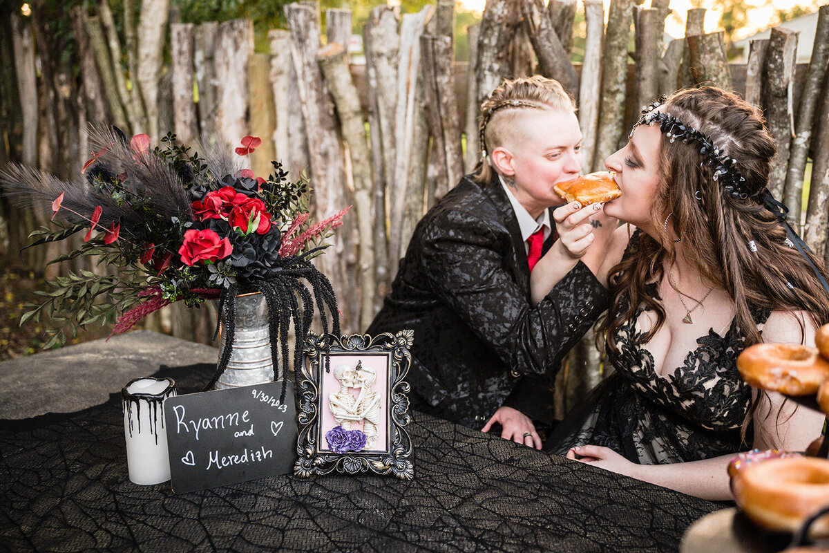 A lesbian couple sits at a cement table with black lace, a pitcher holding a wedding bouquet, a melted candle, a photo with two skeletons kissing, and a chalkboard that reads “Ryanne and Meredith 9/30/2023” as the pair intertwine arms and feed each other donuts from their donut towers.