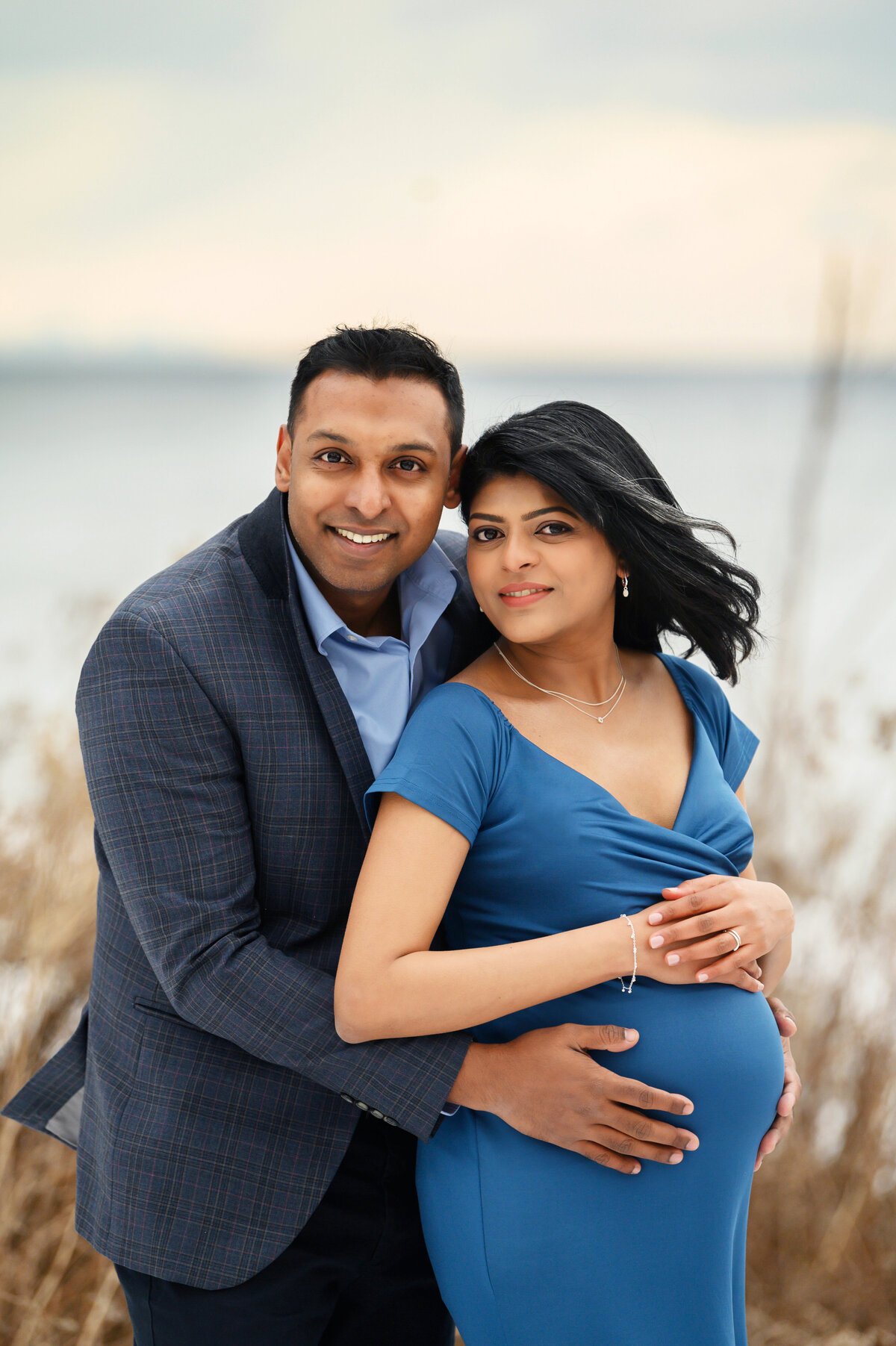Husband standing behind pregnant wife wearing a blue maternity dress holding her belly for their outdoor winter maternity photography session in Burlington, Ontario
