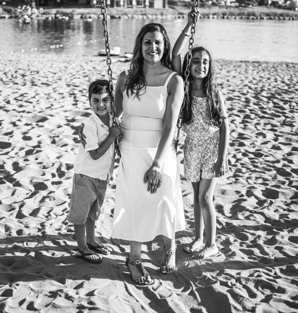 A mom sits on a swing while her son and daughter stand next to her