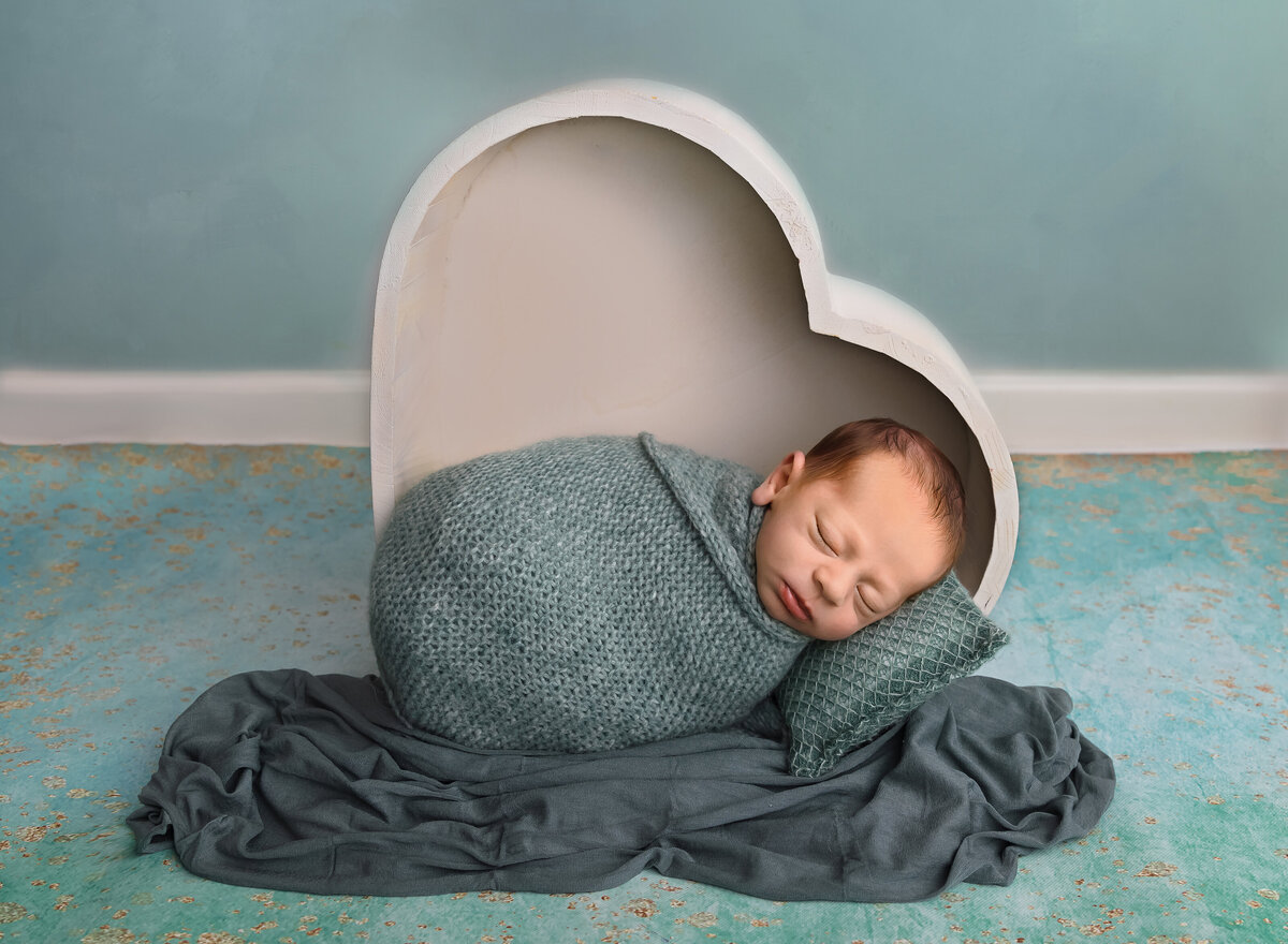 Baby boy is placed in a vertical  heart bowl for a newborn photoshoot. baby is swaddled in deep sage. Captured by top Brooklyn newborn photographer Chaya Bornstein