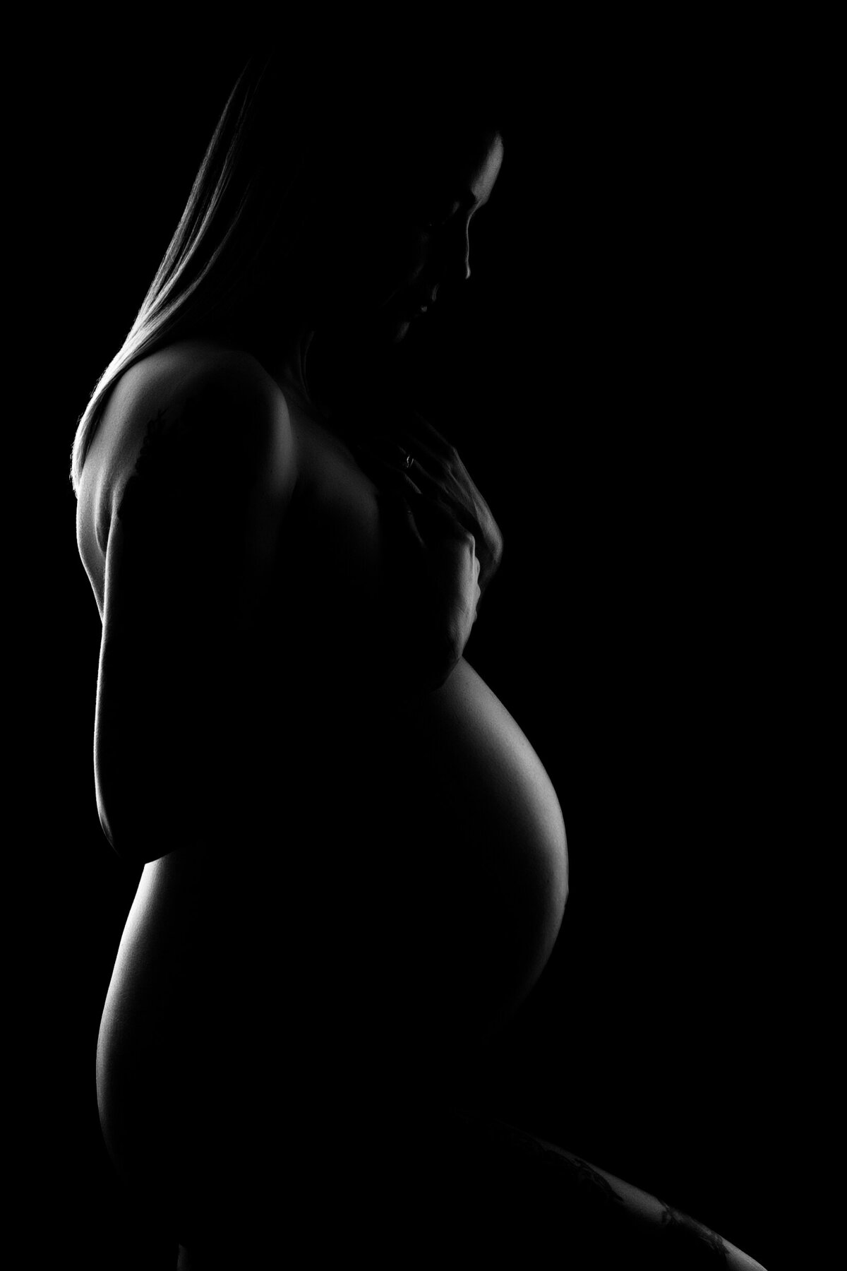 Fine art maternity portrait on a black background with  a little light on the  front and back of the woman's  body.