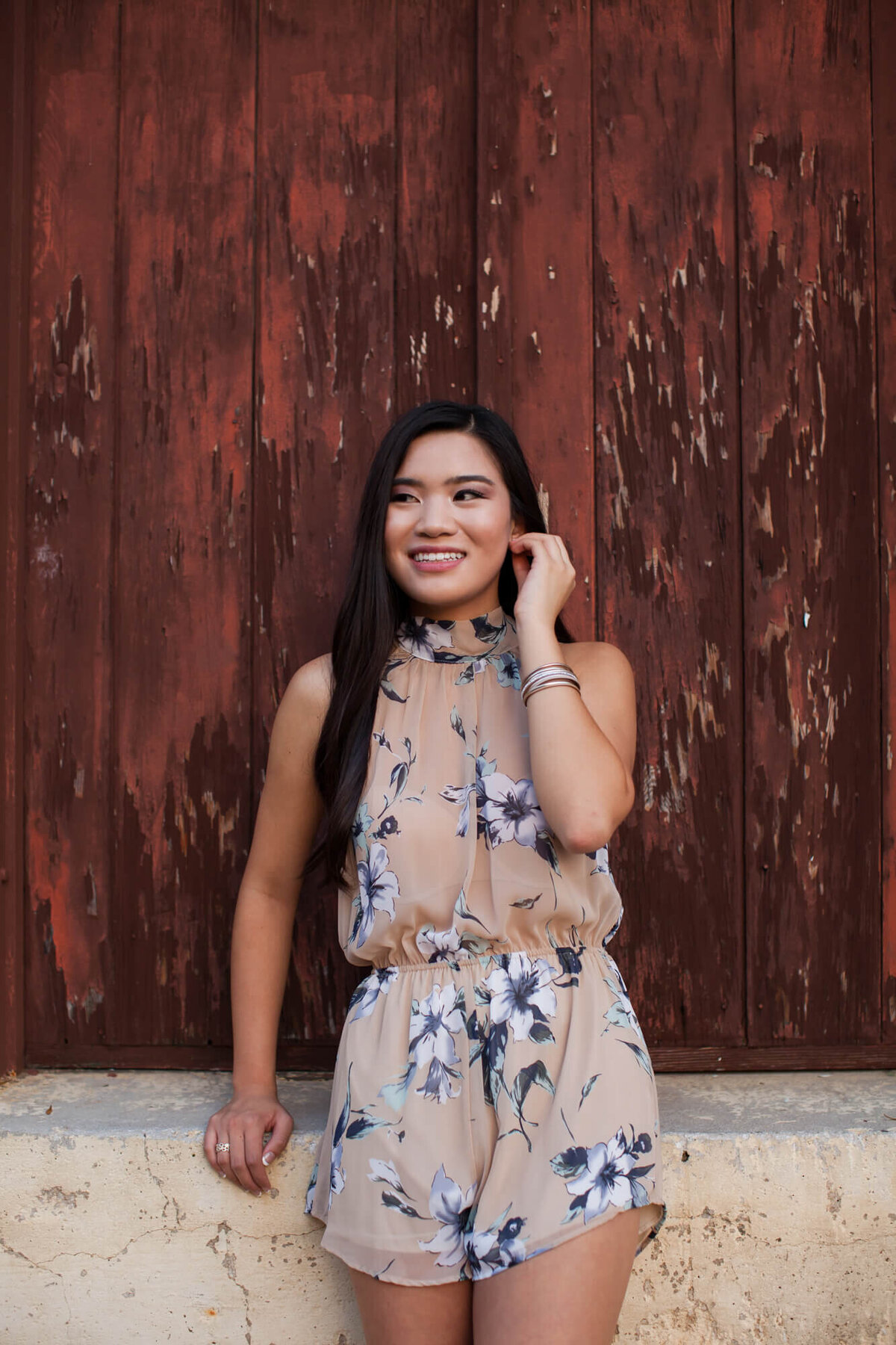 A lovely Asian American girl poses for her senior portraits wearing a floral blush chiffon romper leaning on a window sill with boarded up windows. Captured by Springfield, MO senior photographer Dynae Levingston.