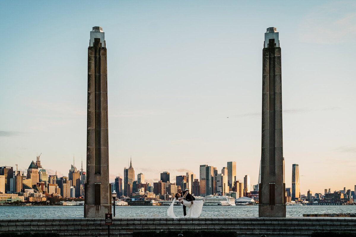 Groom dipping his bride between two tall stone pillars with the Manhattan skyline and Hudson River in the background.