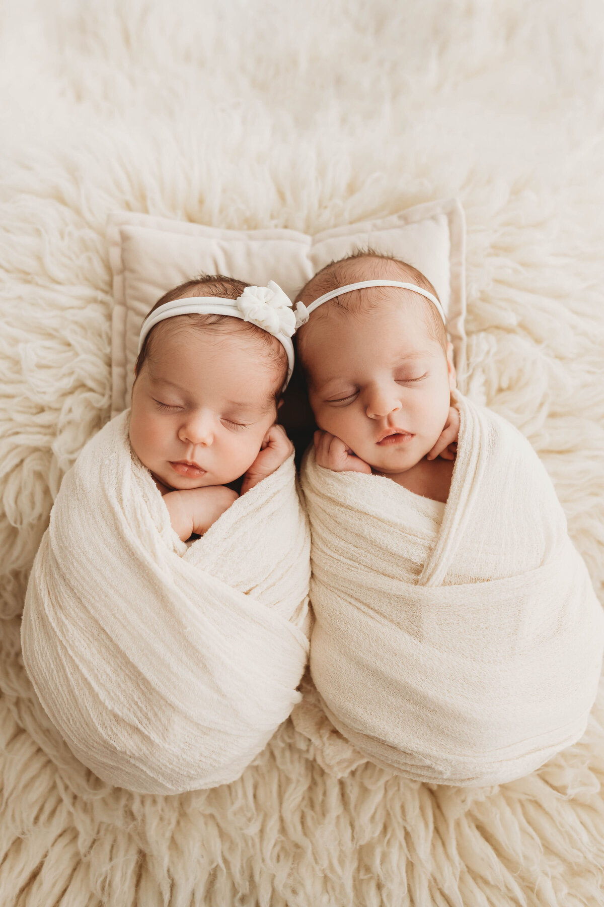 Twin baby girls wrapped in cream swaddles with dainty headbands on.