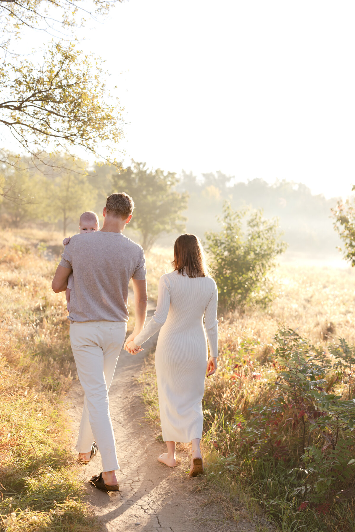a mom and dad hold hands as they walk through a field at sunrise, and they hold htheir baby boy