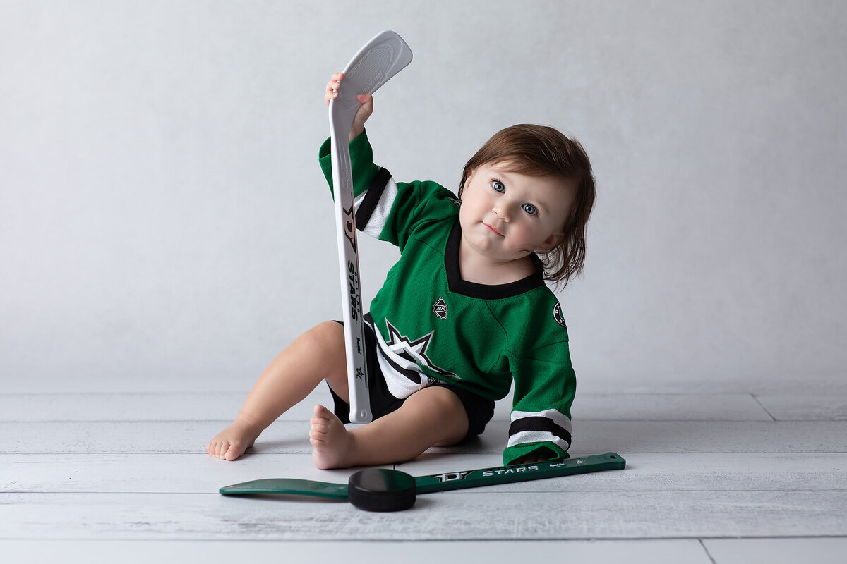 A baby wearing a green Dallas Stars jersey playfully holds a hockey stick while sitting on a white studio floor, showing off their personality during this Dallas baby milestone photographer session.