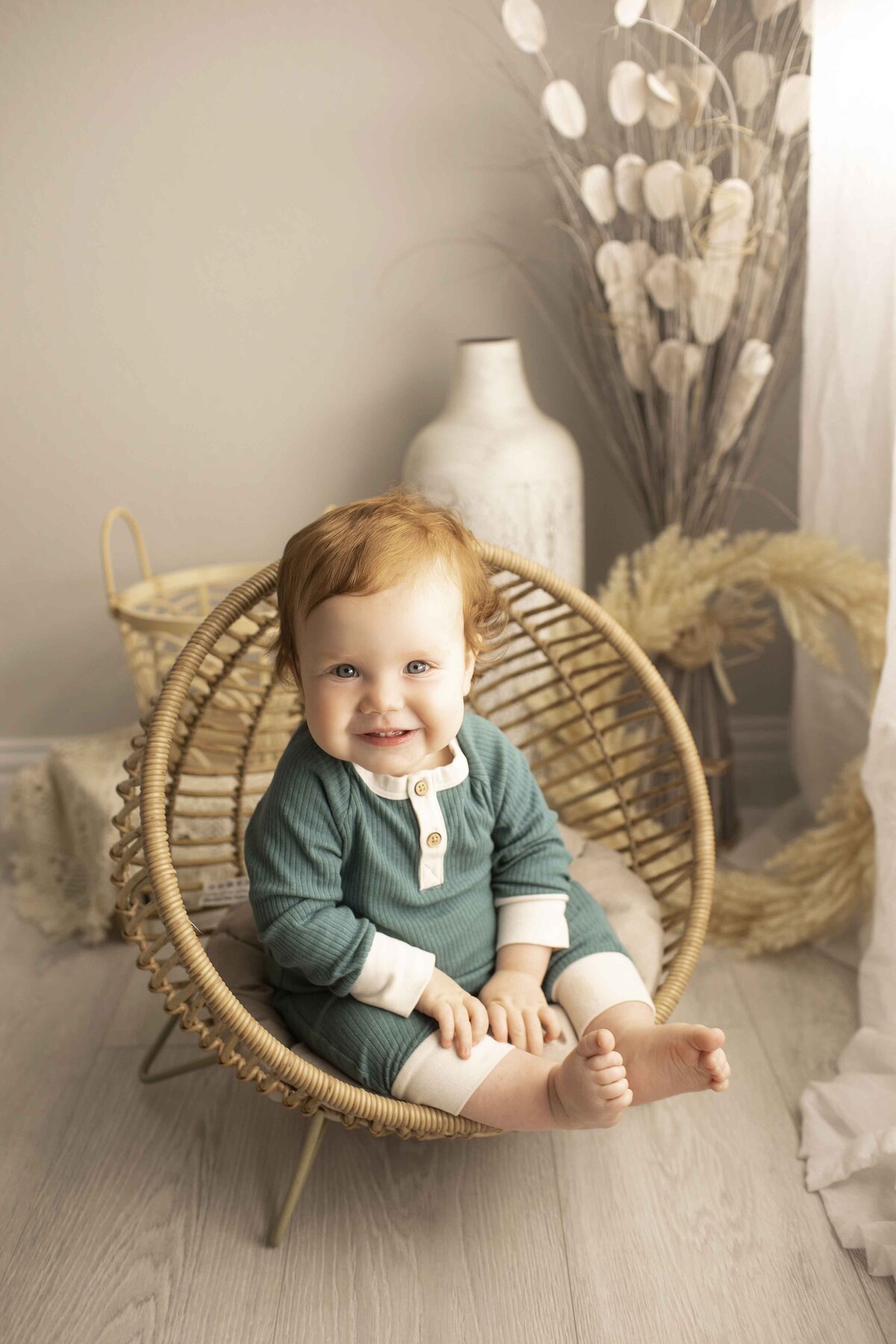 First birthday boy dressed in a cute outfit, sitting in a wicker chair with a big smile in a cozy studio setting
