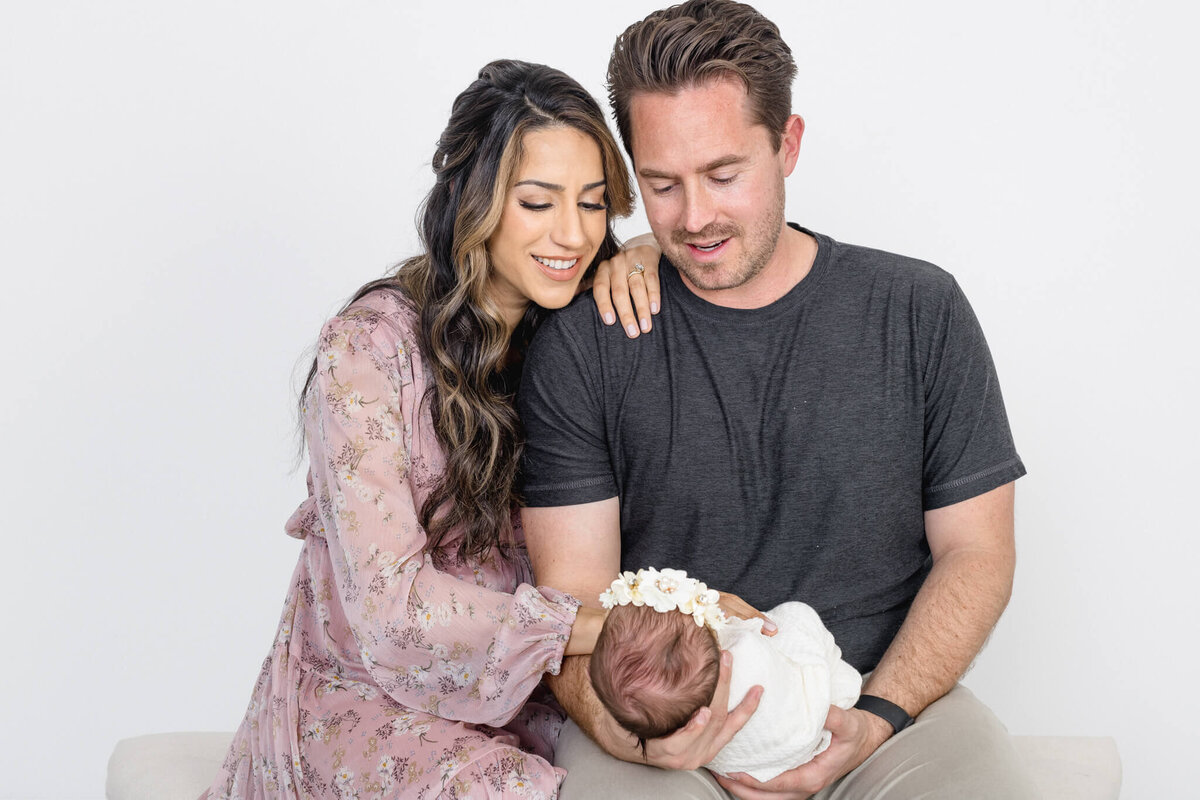 A couple sits together, smiling at a sleeping baby wrapped in a white blanket with a floral headband. The man wears a dark shirt while the woman is in a floral dress.