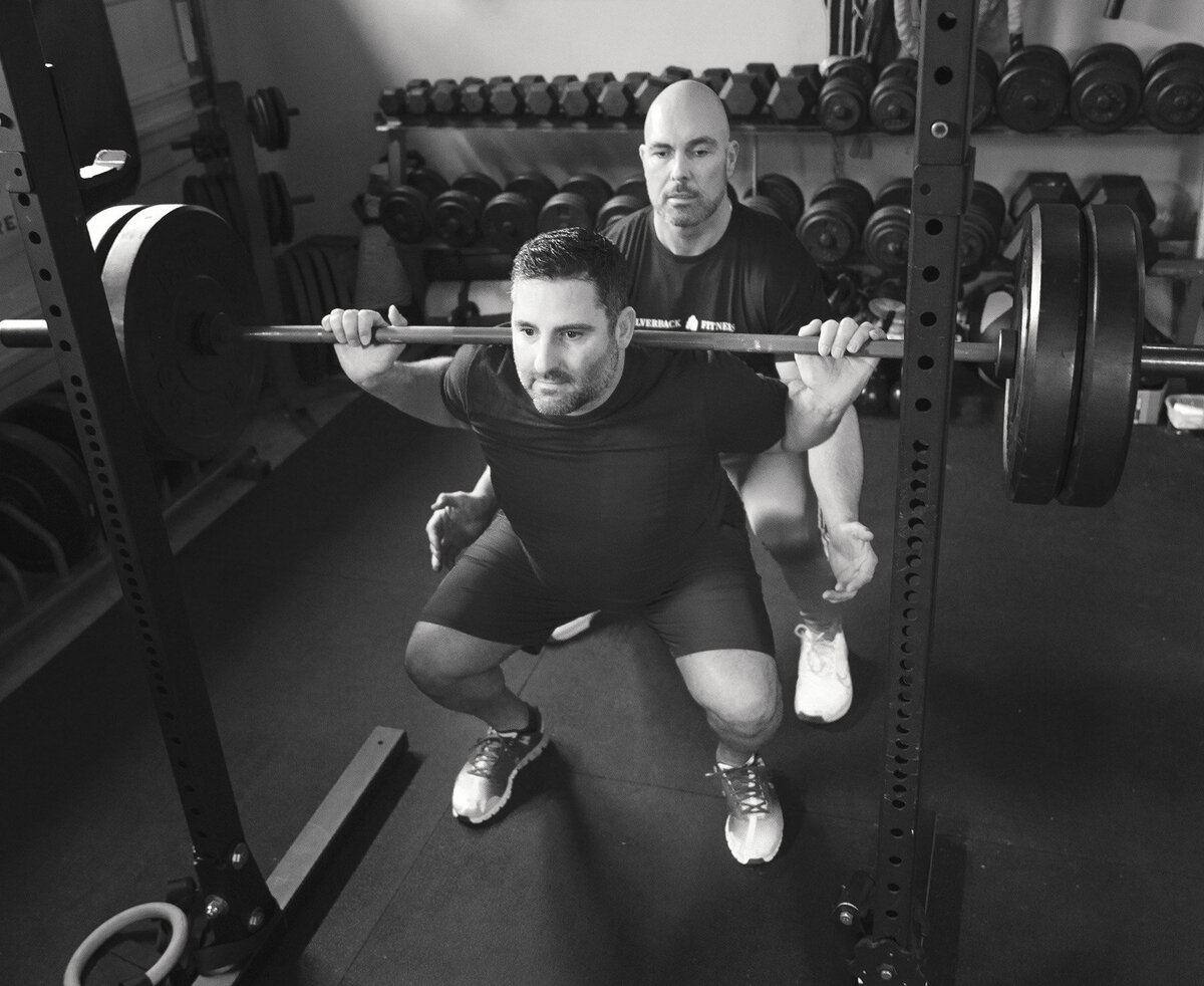 Man is doing squats with barbell at Silverback fitness. He is being spotted by Sarnia's best personal trainer Jim Stewart.