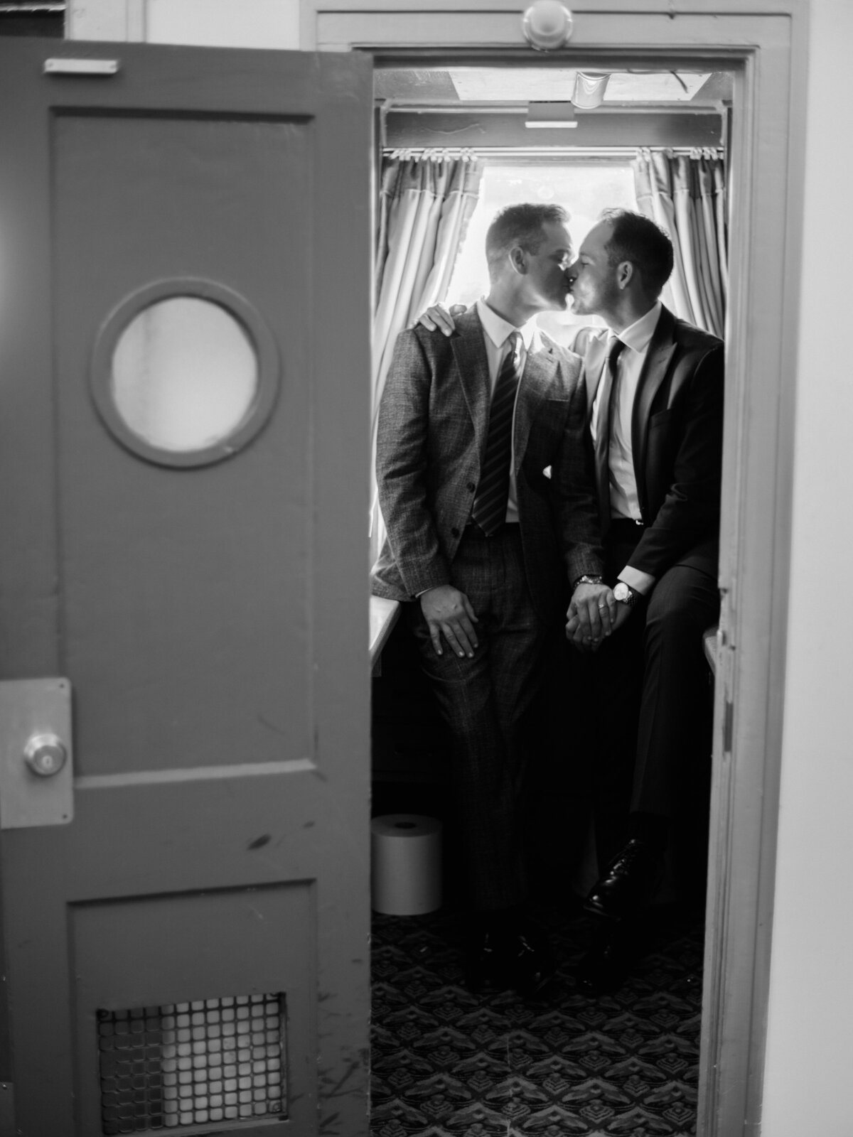 This touching photograph captures the intimate kiss of two suited men in a cozy setting, symbolizing love, tenderness, and connection amidst a quiet backdrop.