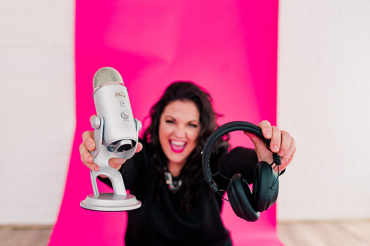 woman sitting on hot pink floor holding up podcast mic and headphones