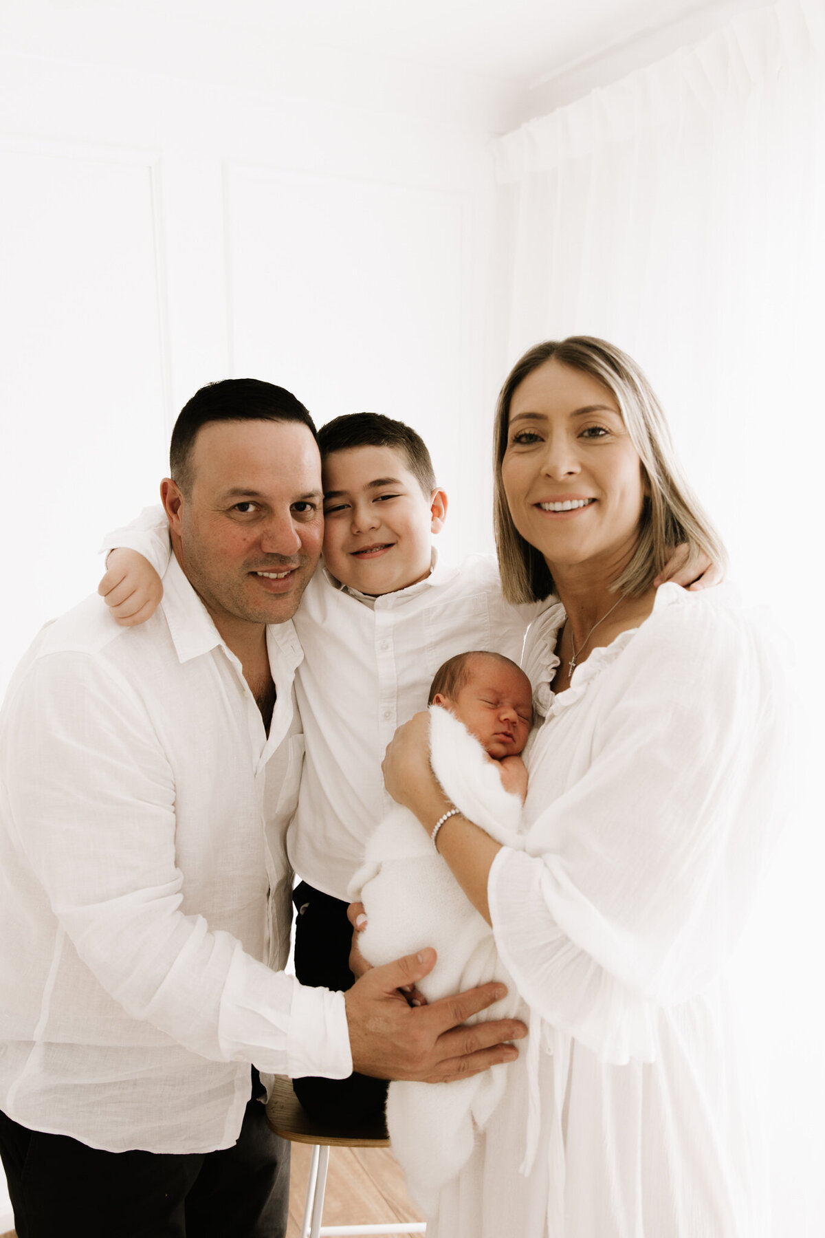 A family of four dressed in white smiling warmly at the camera during a newborn session in a softly lit Southwest Sydney studio, with the mother holding the sleeping newborn.