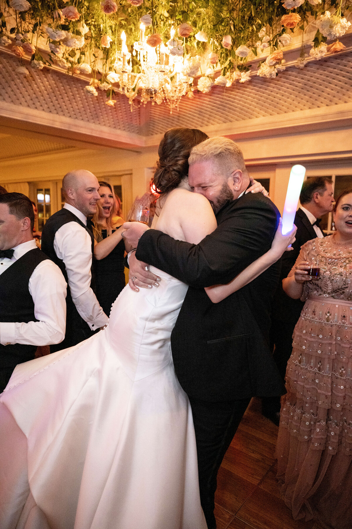 A bride in a white gown embraces a man in a black suit on a dance floor. Guests around them smile and chat, while one holds a glowing stick. A floral chandelier hangs overhead, creating a festive atmosphere.
