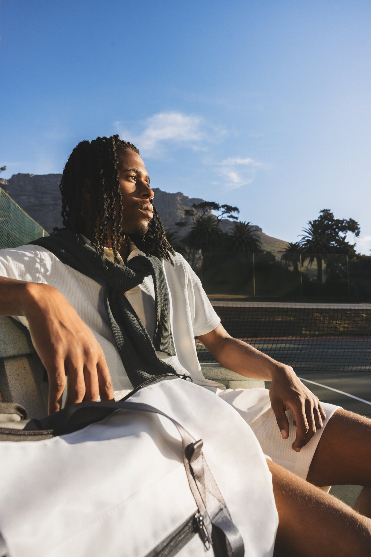 Man in tennis outfit sitting on a bench with a Swim Essentials duffle bag in front, gazing into the distance with palm trees and Table Mountain in the background.