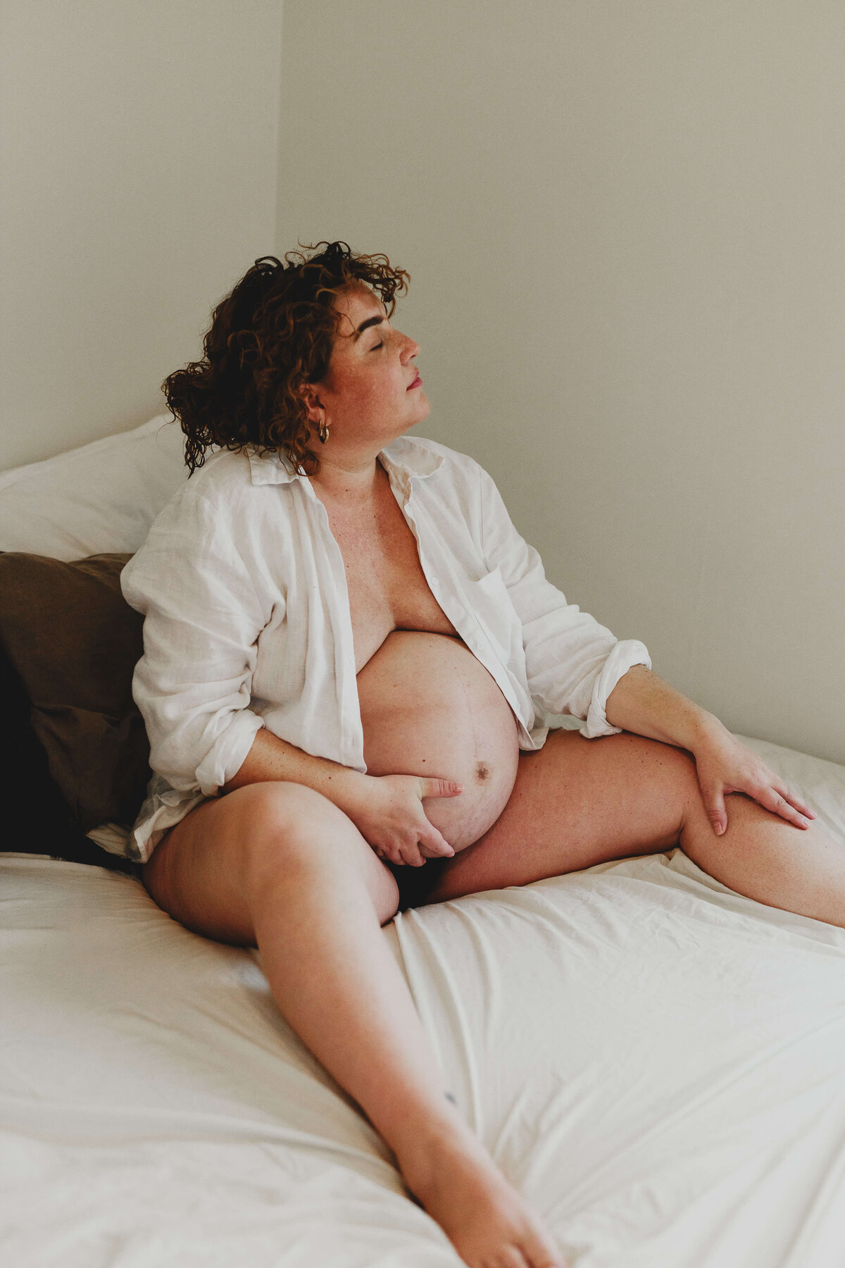 En gravid kvinna sittandes i sin säng med breddade ben tittandes ut mot fönstret under en gravidfotografering i Stockholm