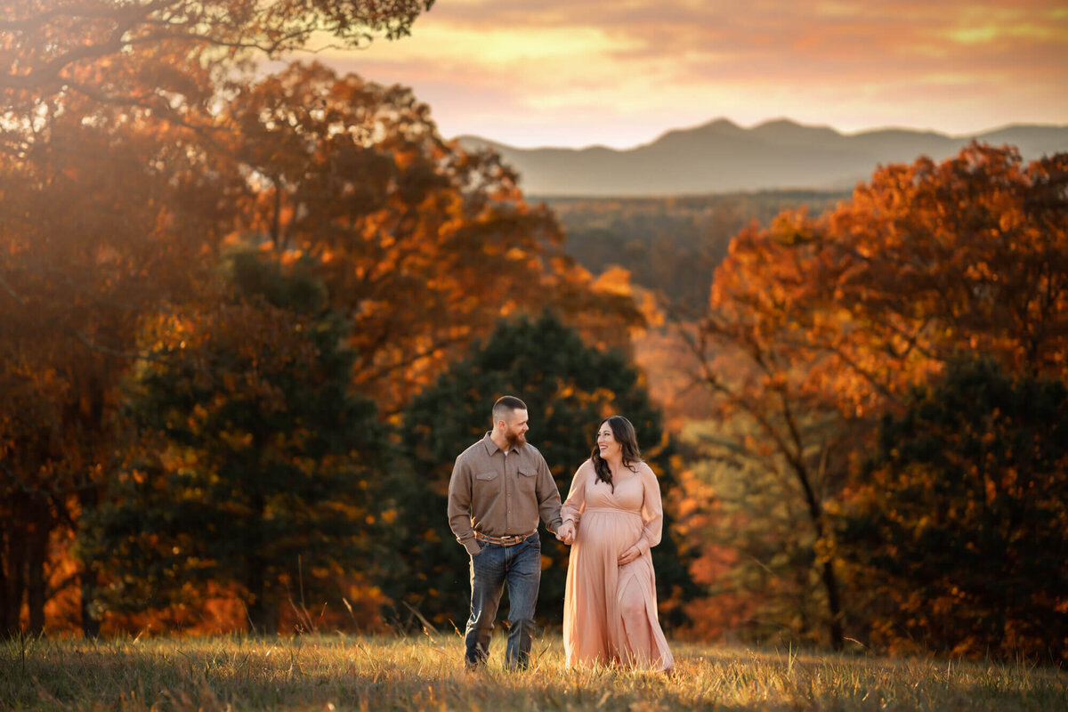 A beautiful young couple expecting their first child holds hands and walks across a meadow as the sun sets.
