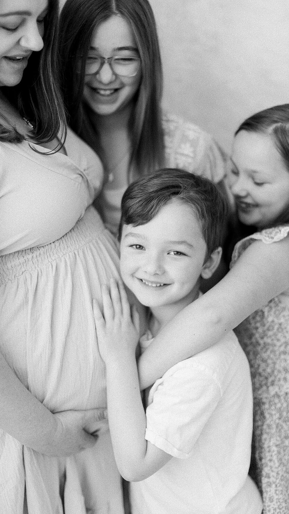 Close up black and white in studio portrait of a mothers growing belly as her two young kids come in for a hug.