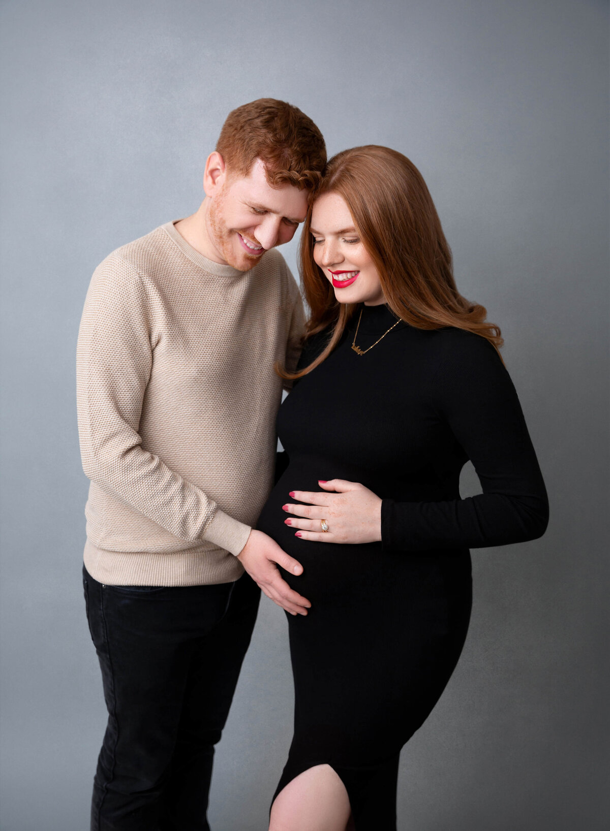 An expecting couple with hands on the bump touch foreheads while standing in a studio in a black dress after meeting a NYC maternity photographer for pregnancy pictures