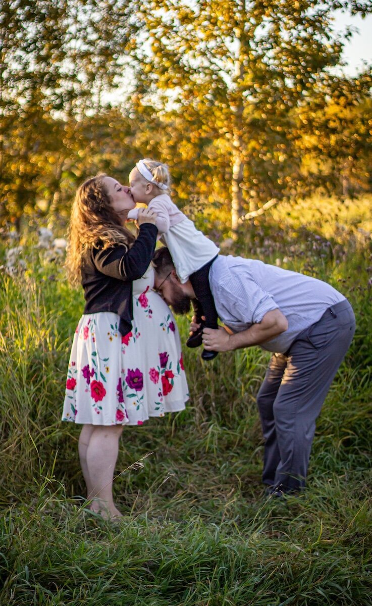 a dad has his toddler daughter on his shoulders leaning forward and kissing his pregnant wife's belly