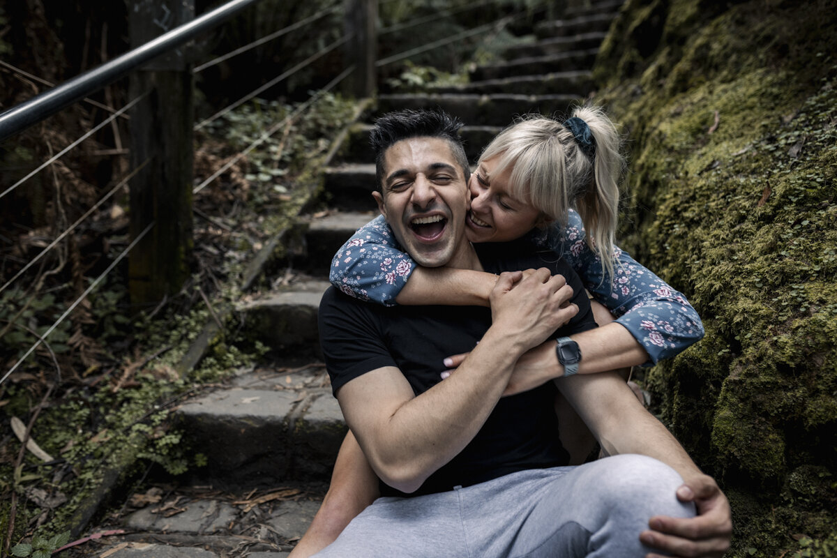 Bride and Groom standing on a mountain top. The wind is blowing in her hair and they are looking out to the view while hugging each other. The bride is a long sleeved wedding gown.