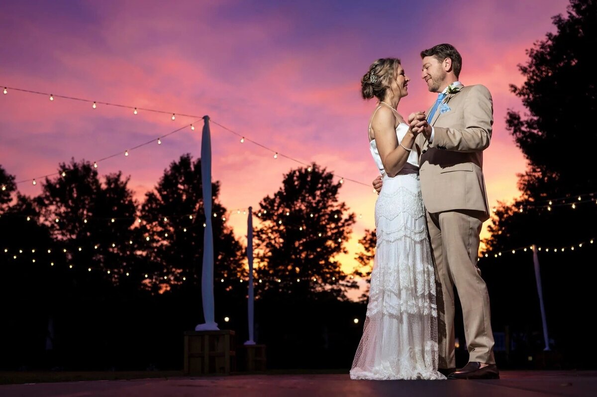 A bride and groom share a dance under a twilight sky, with string lights above and a vivid sunset in the background, creating a romantic and picturesque scene.