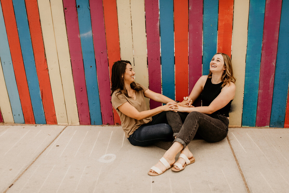 lesbian couple sitting and laughing in front of a rainbow fence