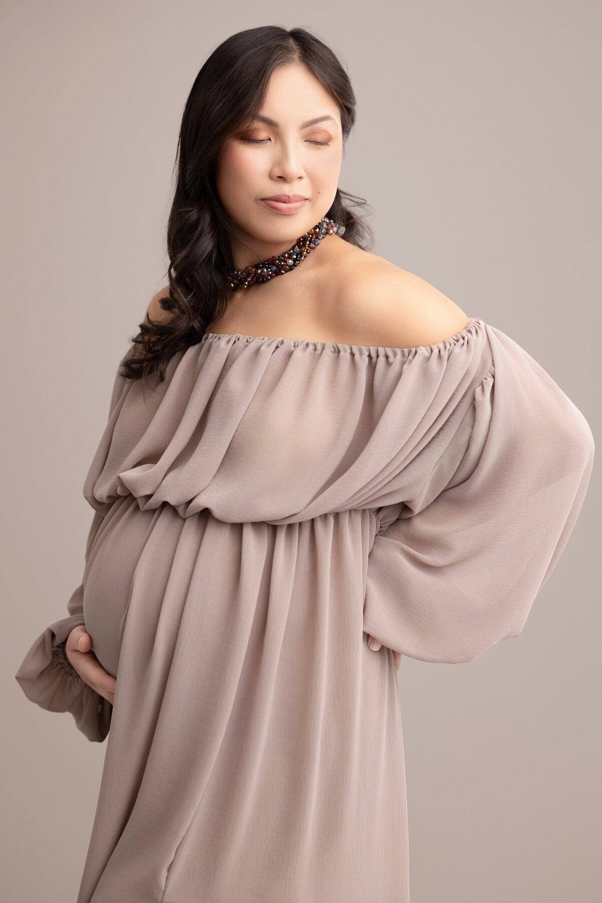 Pregnant mother wearing a brown off-the-shoulder dress and multi-colored necklace, with one hand under belly and the other hand on hip.