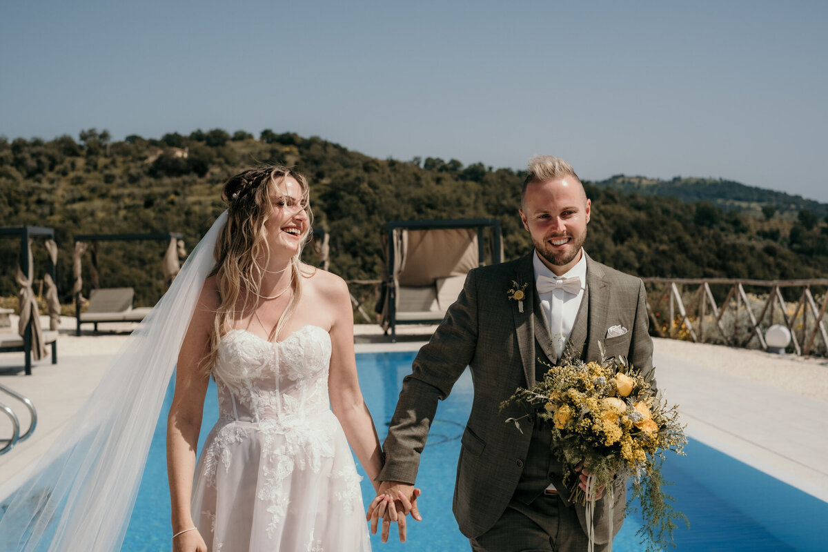 Das Hochzeitspaar lacht entspannt, als es vor einem Pool und Hügeln im Hintergrund entlang läuft.