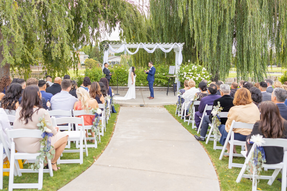 Wedding ceremony with bride and groom reading their vows to each other. Photo is taken from center of aisle with guests looking on.  Captured by Sacramento photographer studio, Philippe Studio Pro