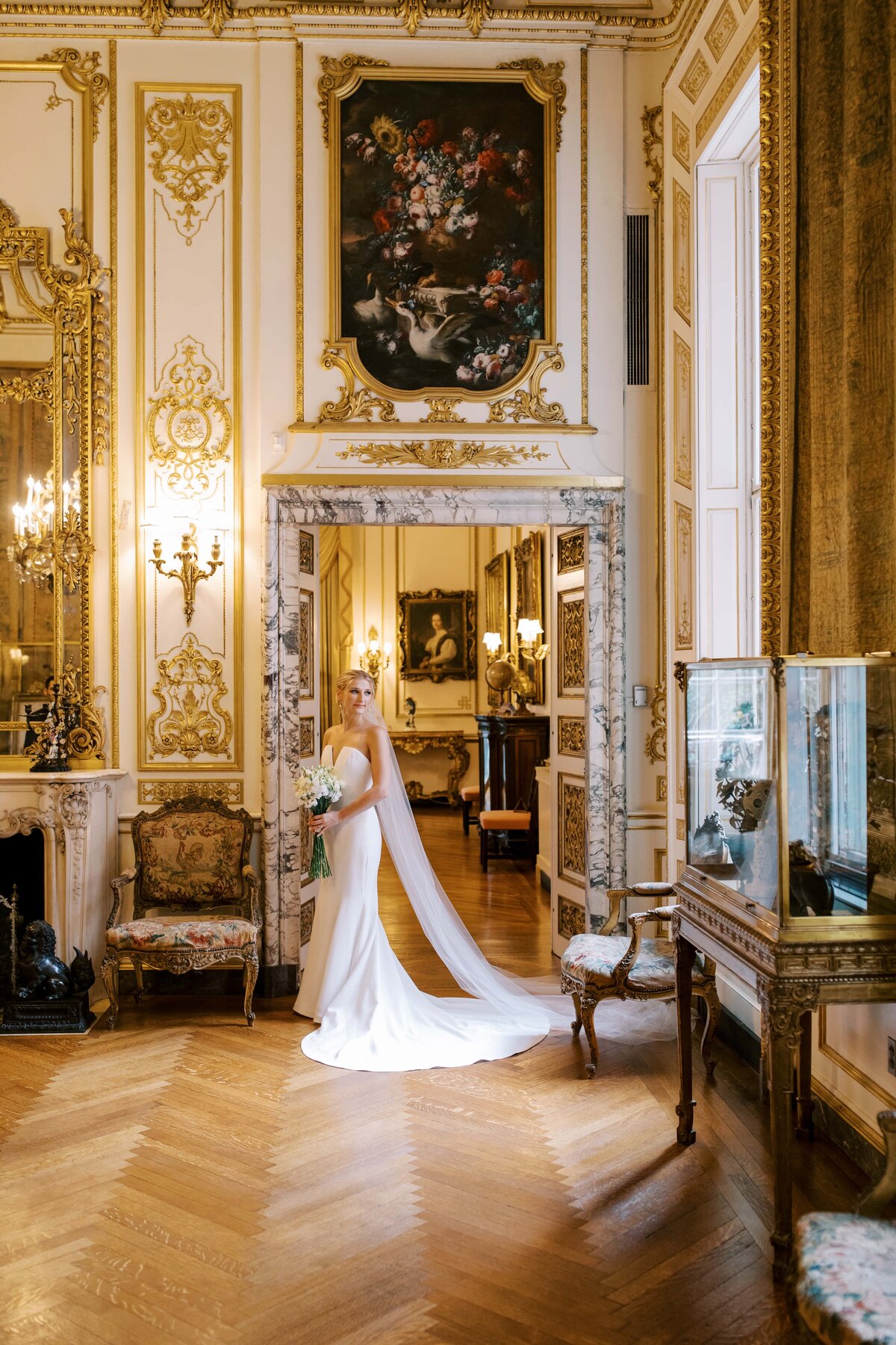 A fair haired bride in a white wedding gown holding a bouquet of white flowers leaning against a wall in an ornately decorated gold room in the Anderson House in Washington D.C.